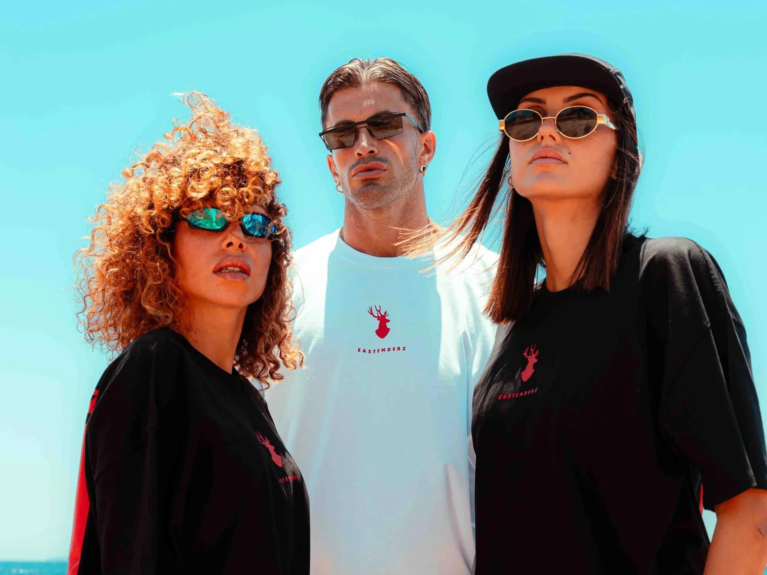 Three young adults stand close together against a blue sky, wearing sunglasses and casual black and white clothing with a red logo, looking confidently at the camera.