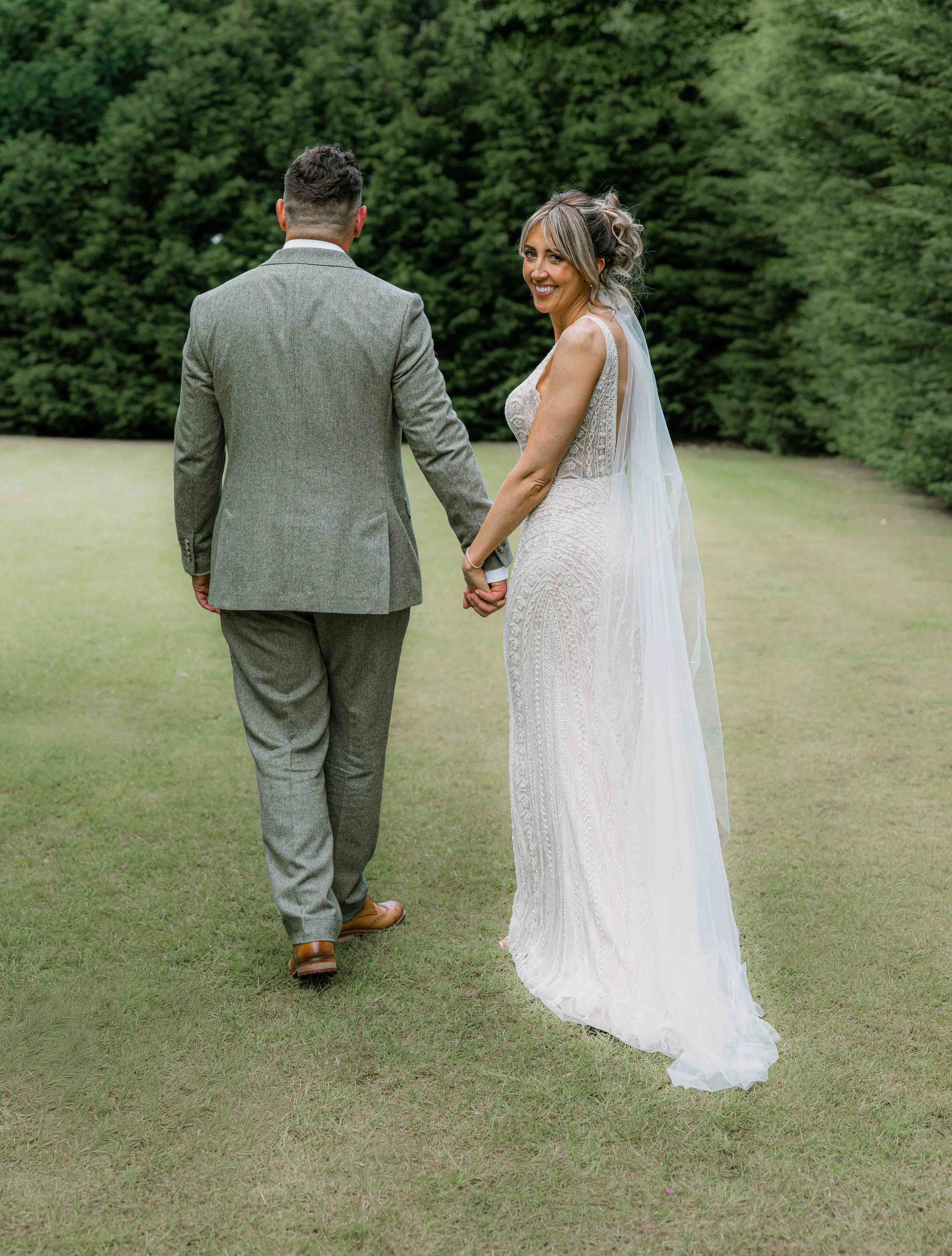 A bride and groom holding hands outdoors on a grassy area with trees in the background, smiling and looking at each other.