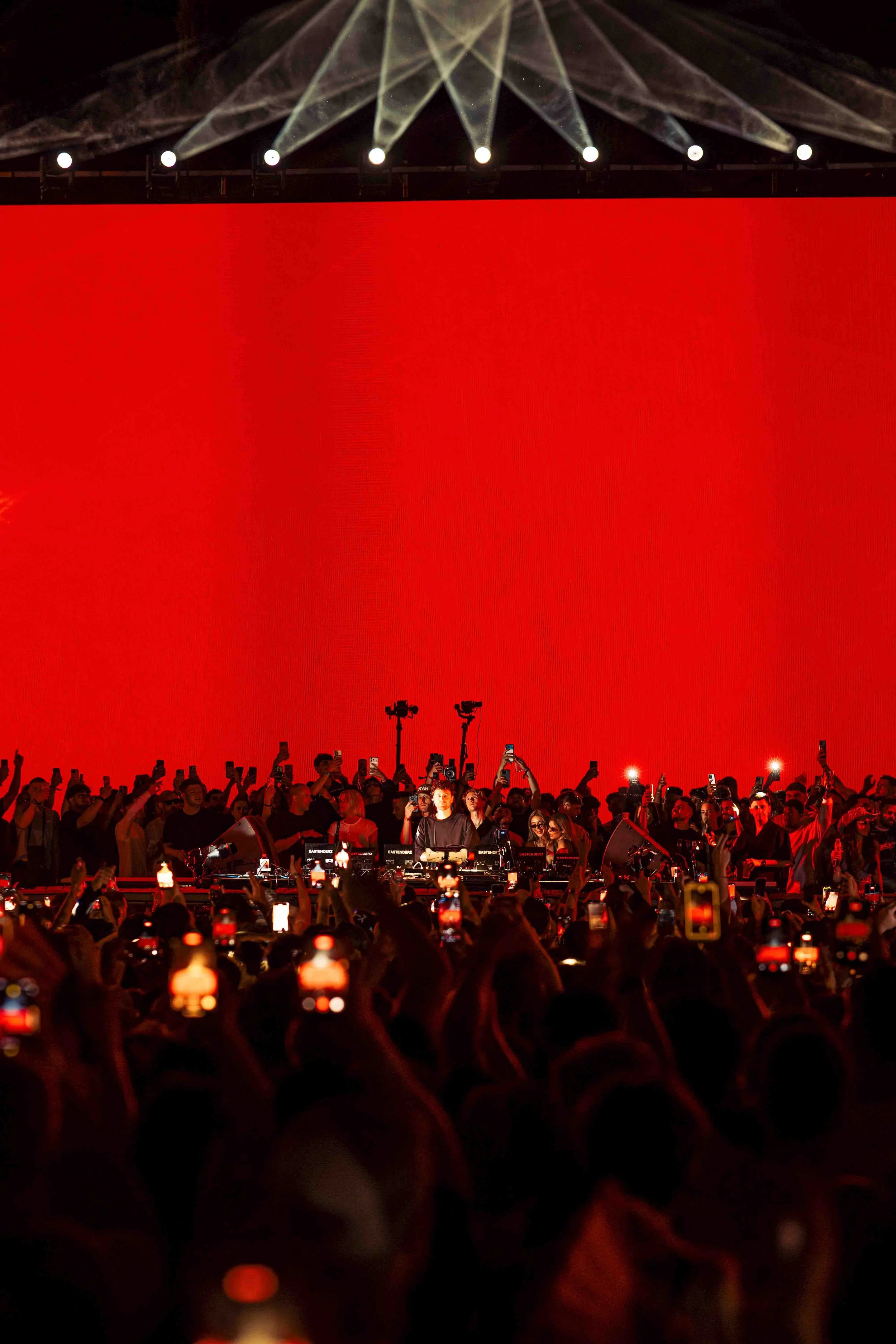 A DJ performing at a concert with a large crowd gathered in front, many holding up phones to record. The background features a large bright red screen and bright stage lights shining down.
