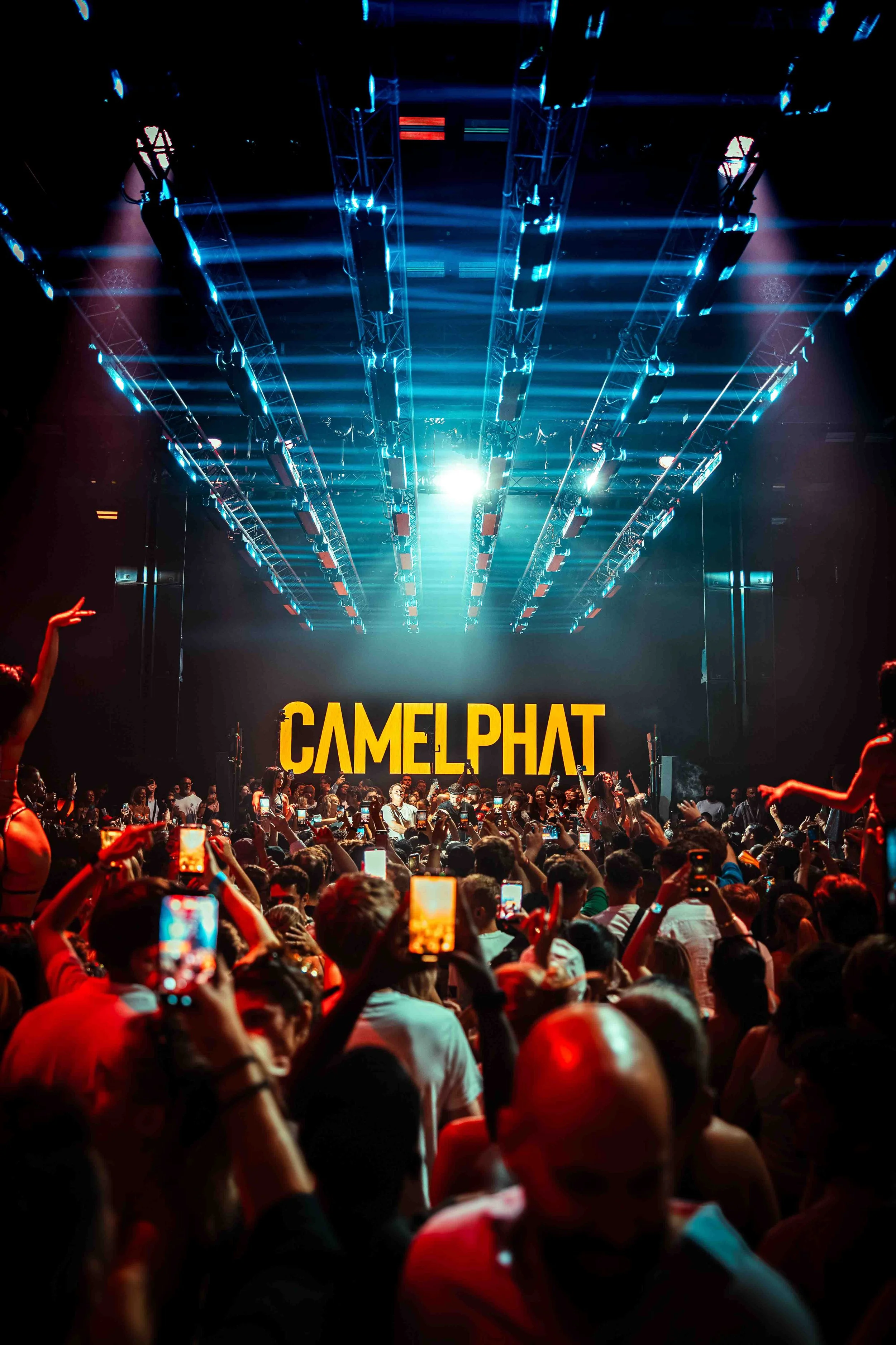 Crowd of people at a concert or event with a large stage displaying the words "CAMELOT" in bold yellow letters, and bright blue stage lights illuminating the scene.