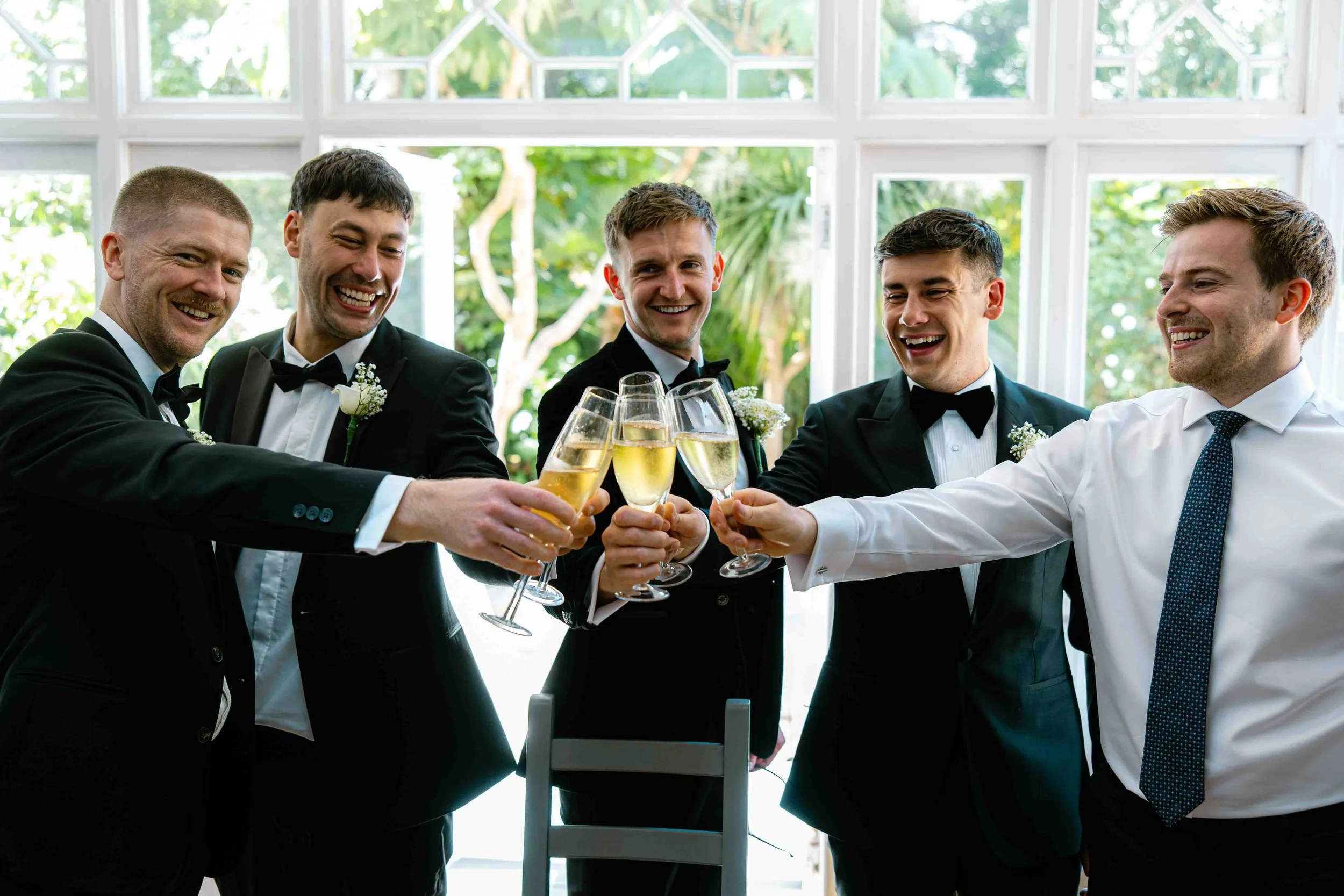 A group of five men in tuxedos clinking glasses of champagne during a celebration indoors with large windows showing green trees outside.