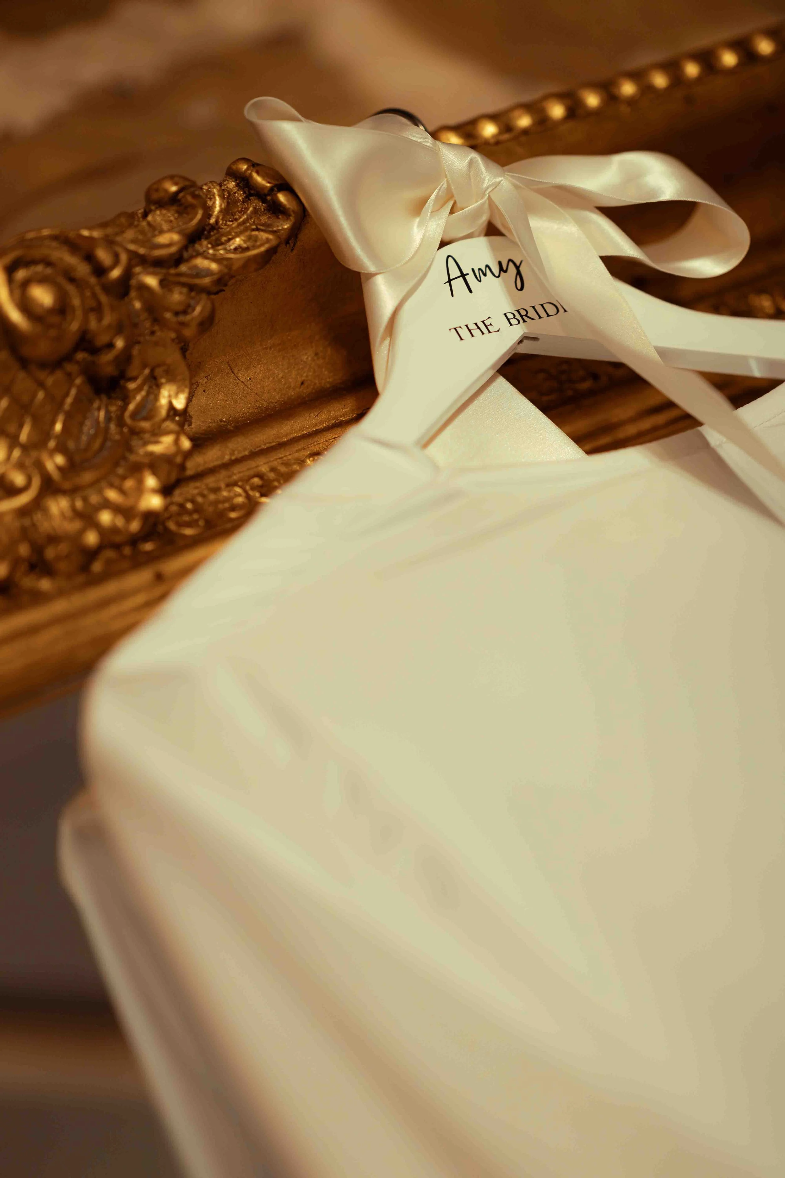 A decorative gold-framed mirror with a white satin ribbon tied around it and a tag that reads 'Amy THE BRIDE' hanging from the ribbon.
