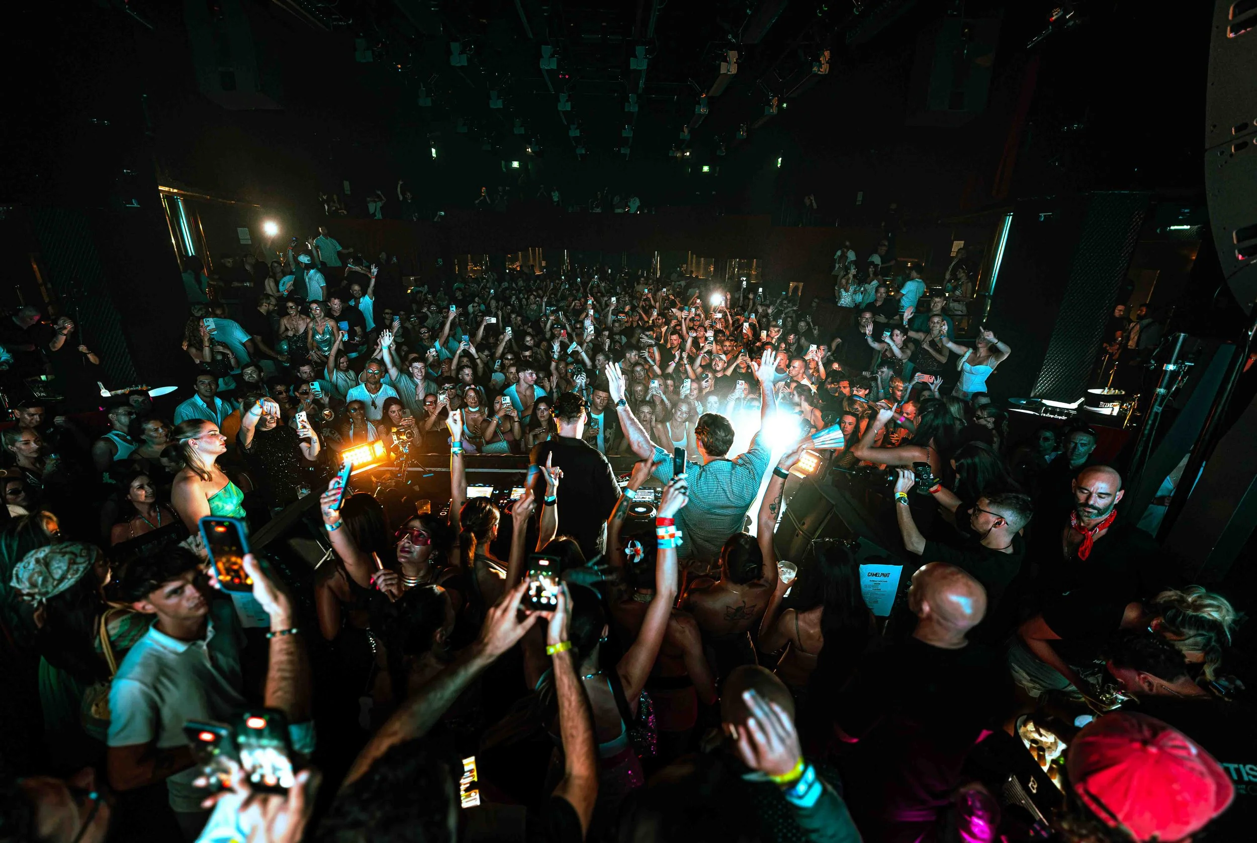Crowd of people at a nightclub or concert, many holding up smartphones, with DJ in the foreground and stage lighting.