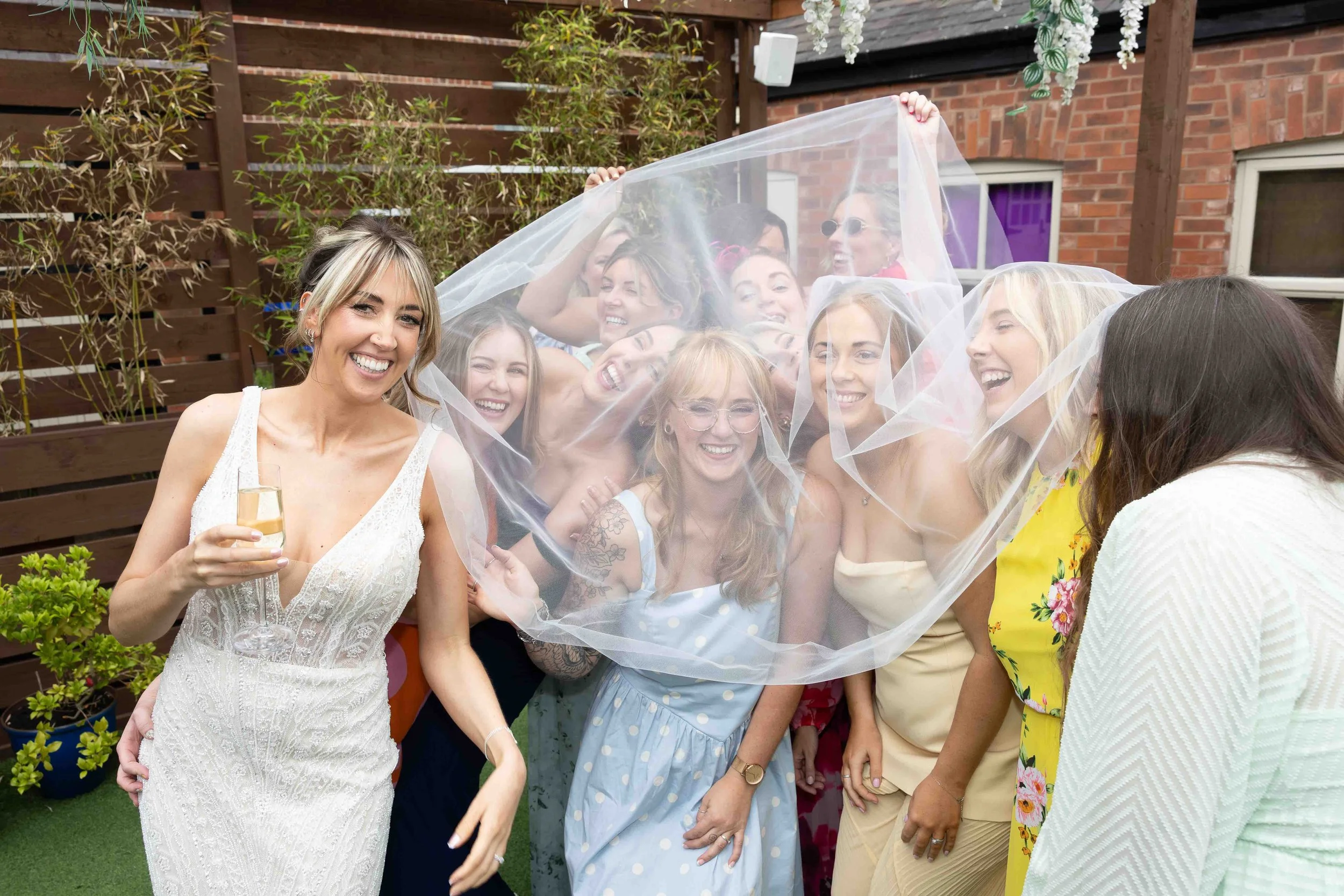 A group of women laughing and celebrating outdoors, with one woman in a white dress holding a glass of champagne. Others are gathered under a sheer veil, enjoying the moment.