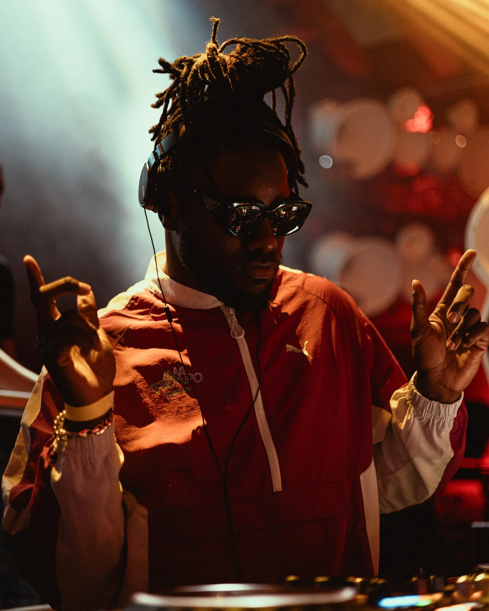 A person with dreadlocks wearing sunglasses, headphones, and a red jacket with white sleeves and a zipper, holding DJ equipment. The background has bokeh lights.