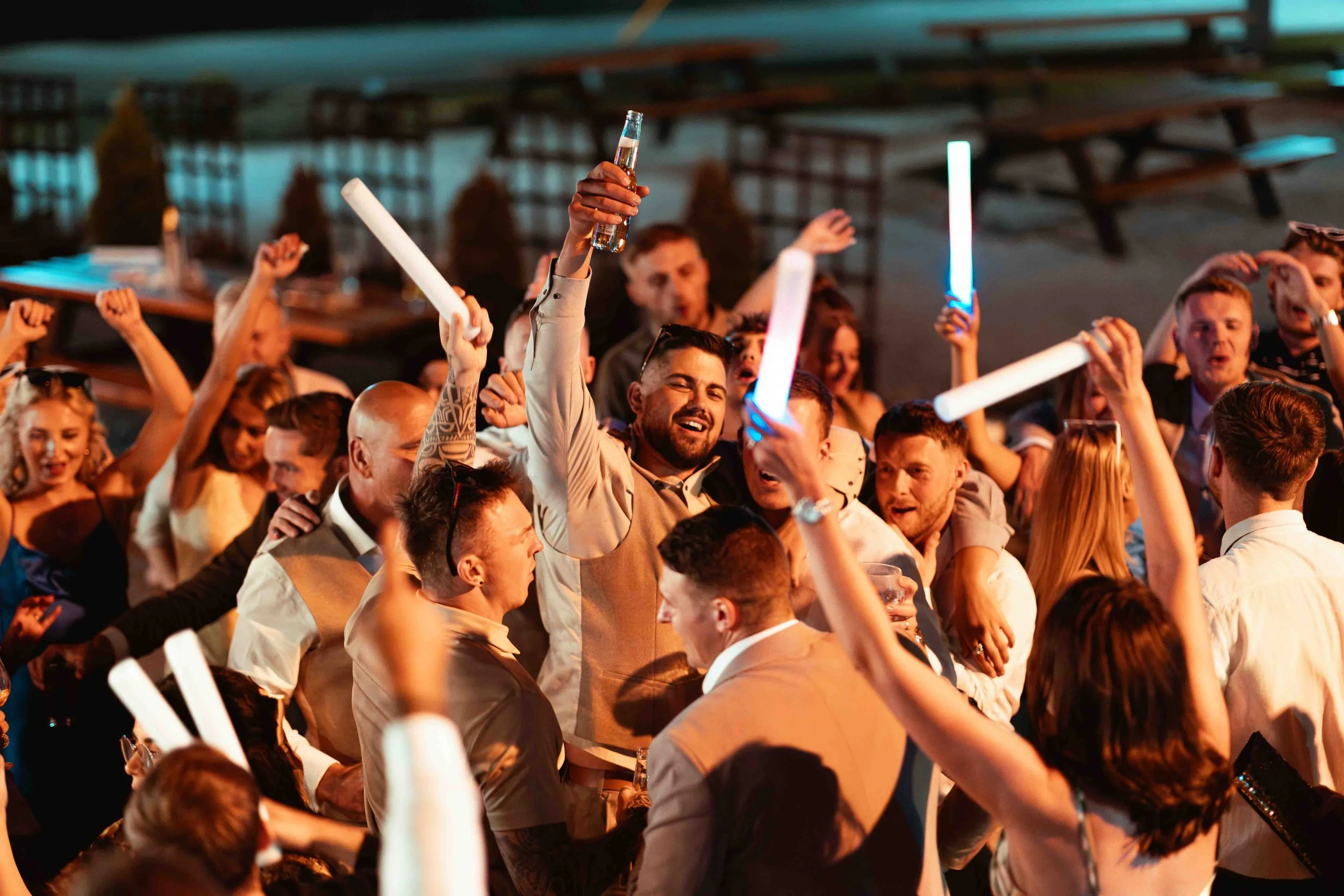 A crowded party or celebration with many people holding glow sticks, some raising drinks, in a dimly lit indoor setting.