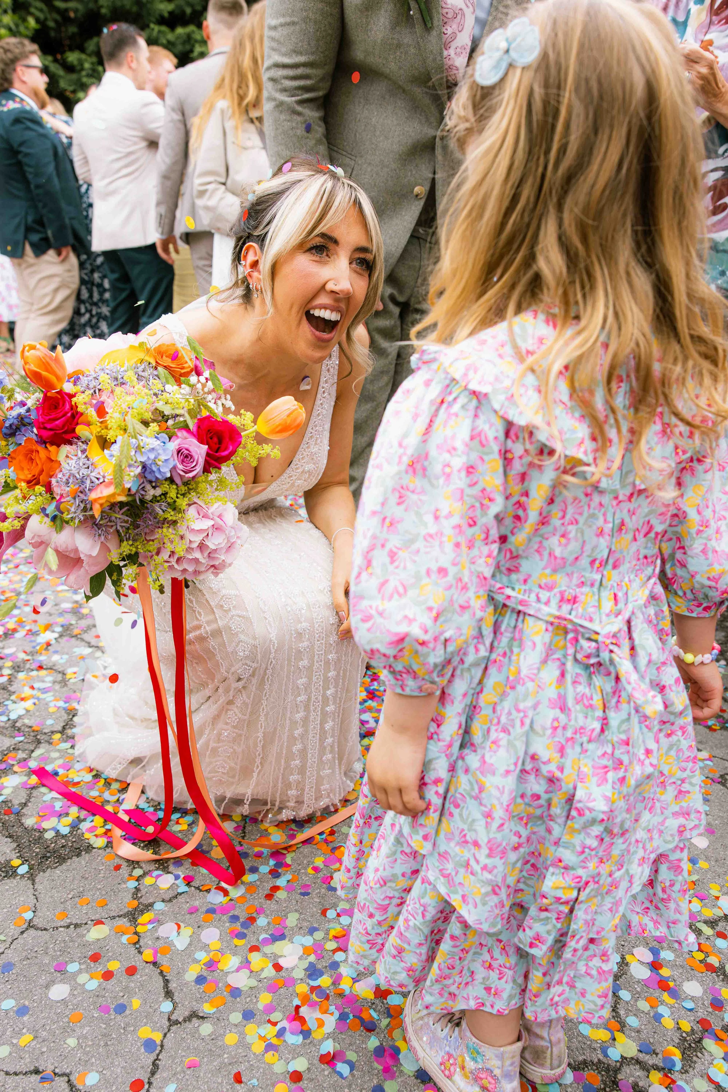 A woman in a wedding dress holding a colorful bouquet, kneeling on the ground surrounded by multicolored confetti, with a joyful expression, during a celebration or wedding celebration.