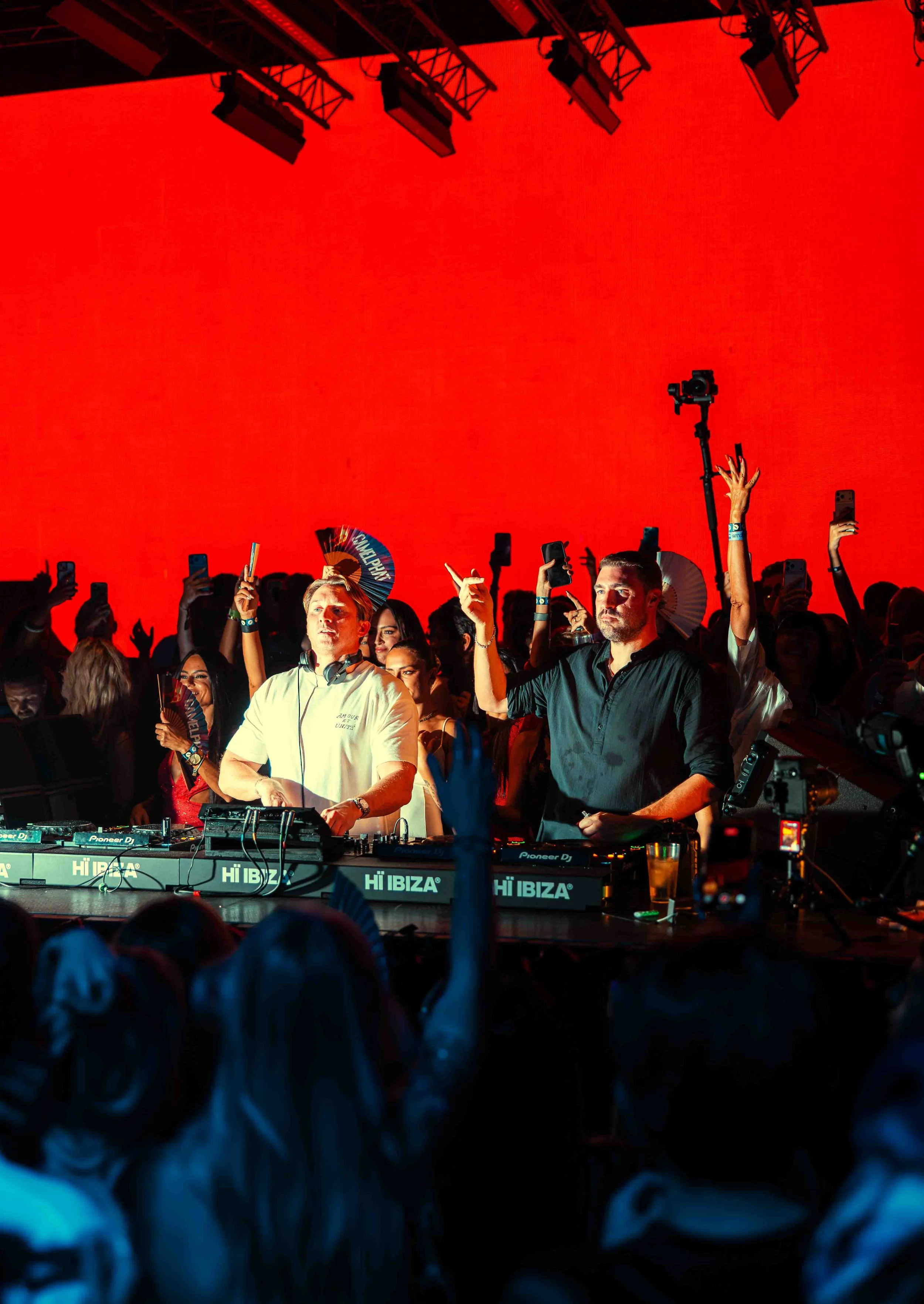 Crowd at a concert or festival, with a DJ performing on stage with a large red backdrop and people taking photos or videos with their smartphones.