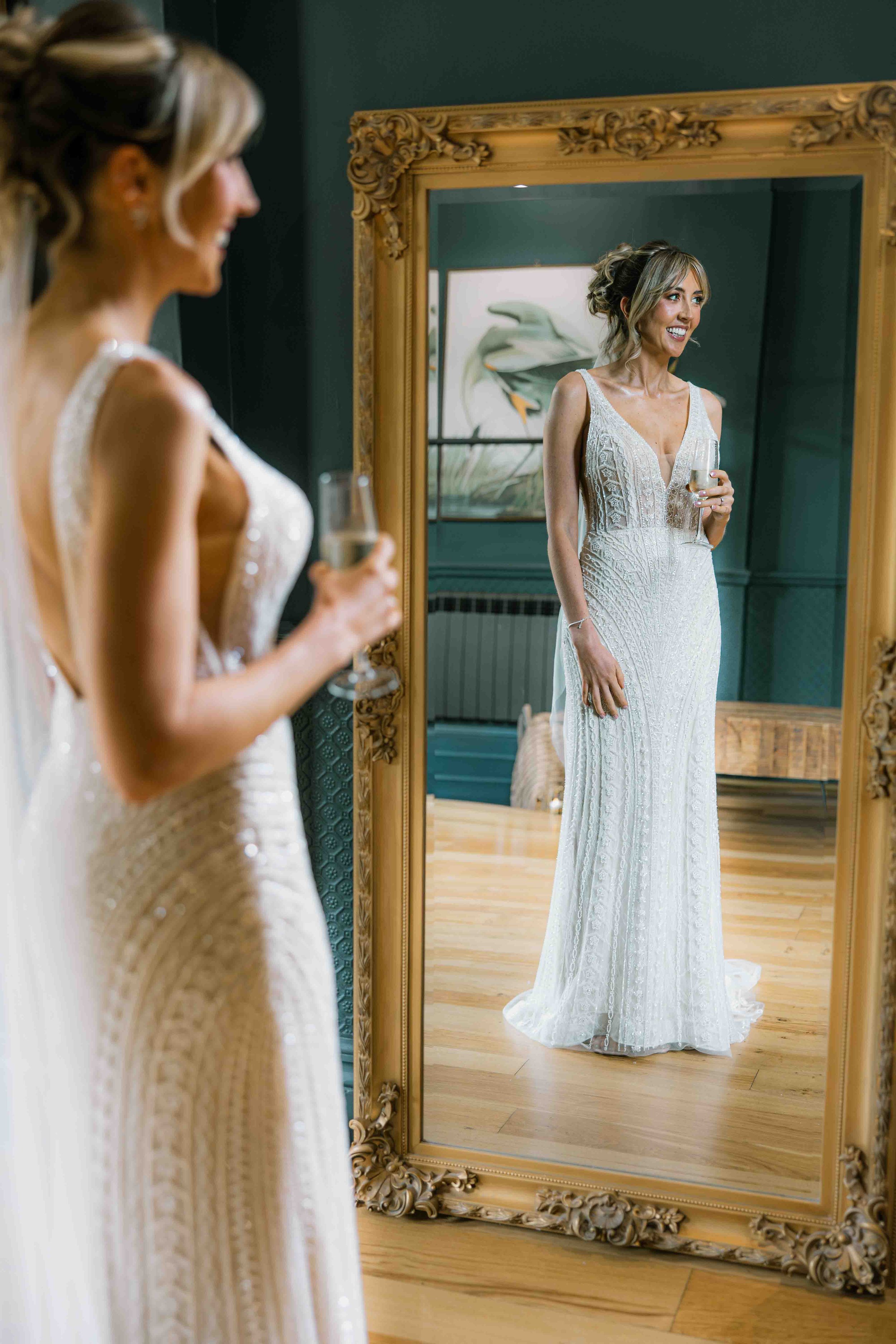 A woman in a wedding dress smiling at her reflection in a large, ornate mirror, holding a glass of champagne.