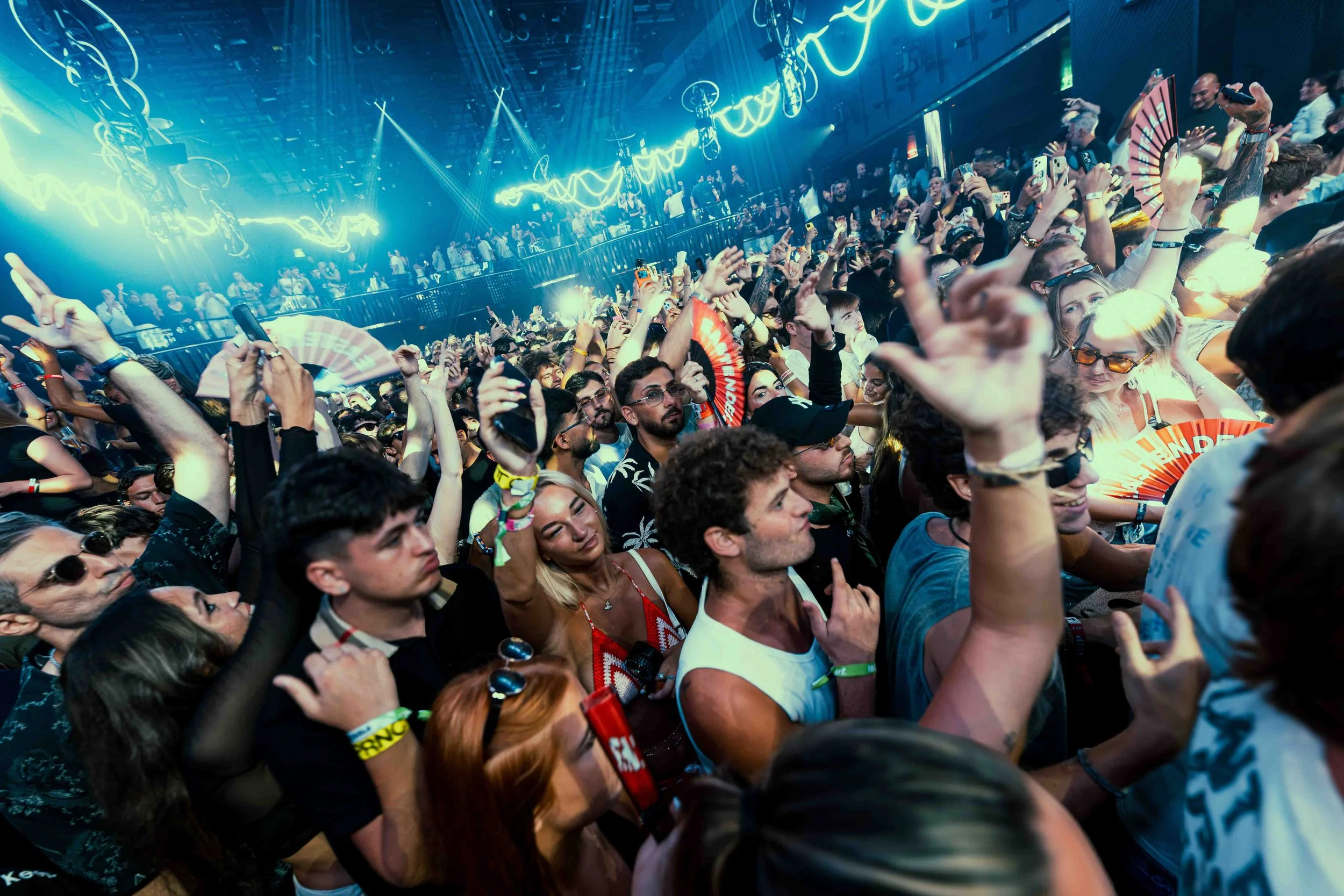 Crowd of people at a concert or music event with neon lights and people holding up phones and fans.