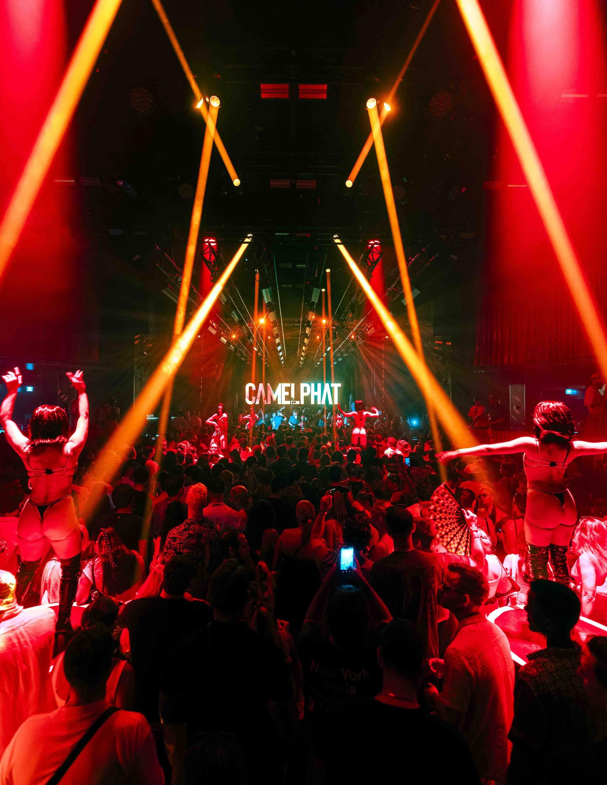 A crowded nightclub with people dancing and enjoying a live performance or DJ. The stage is illuminated with colorful and bright lighting, including yellow and red beams. A large sign displaying the word 'CAMP PHAT' is visible at the back of the stag