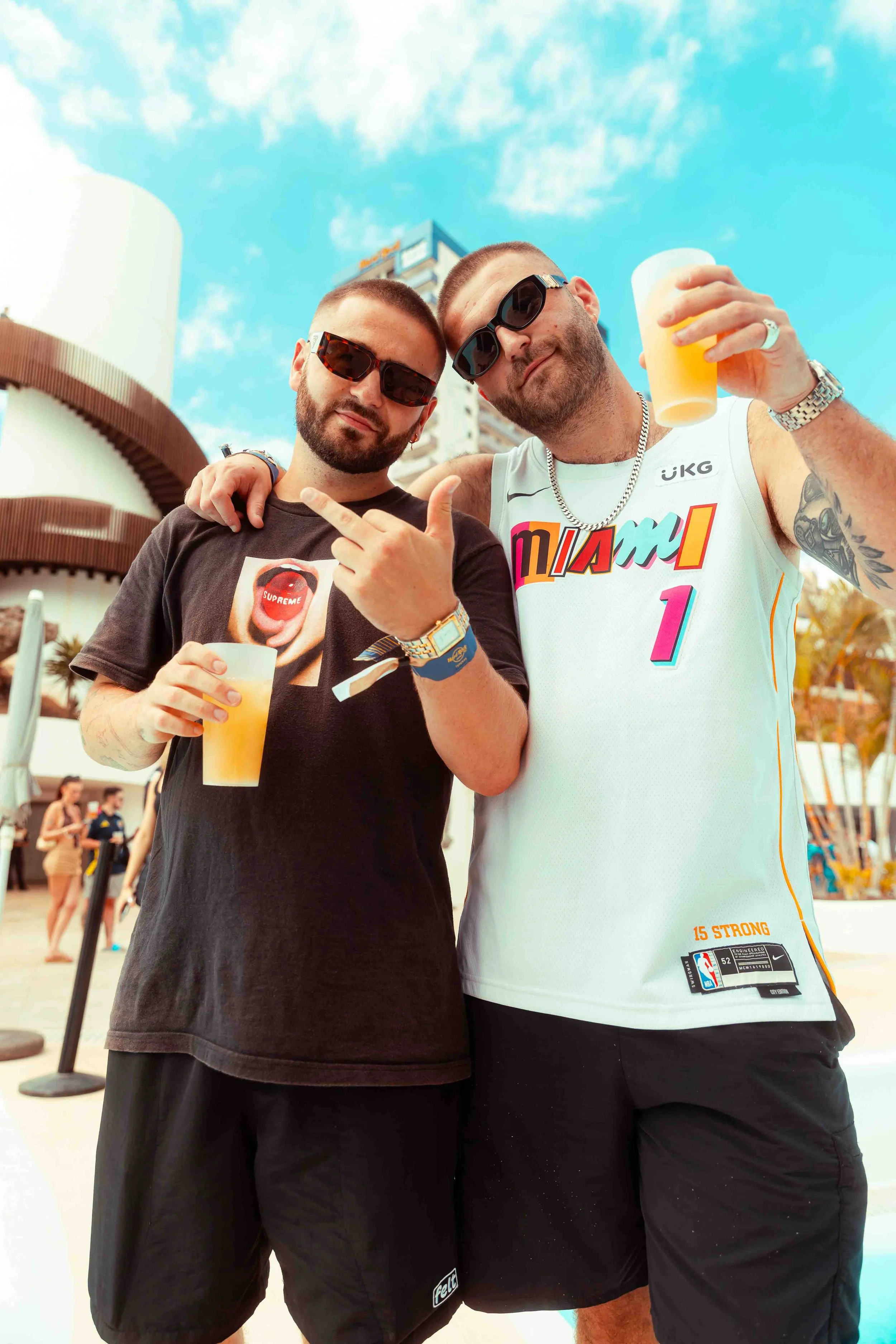 Two men standing close together outdoors, holding drinks, wearing sunglasses, and smiling at the camera, with a sky and modern buildings in the background.
