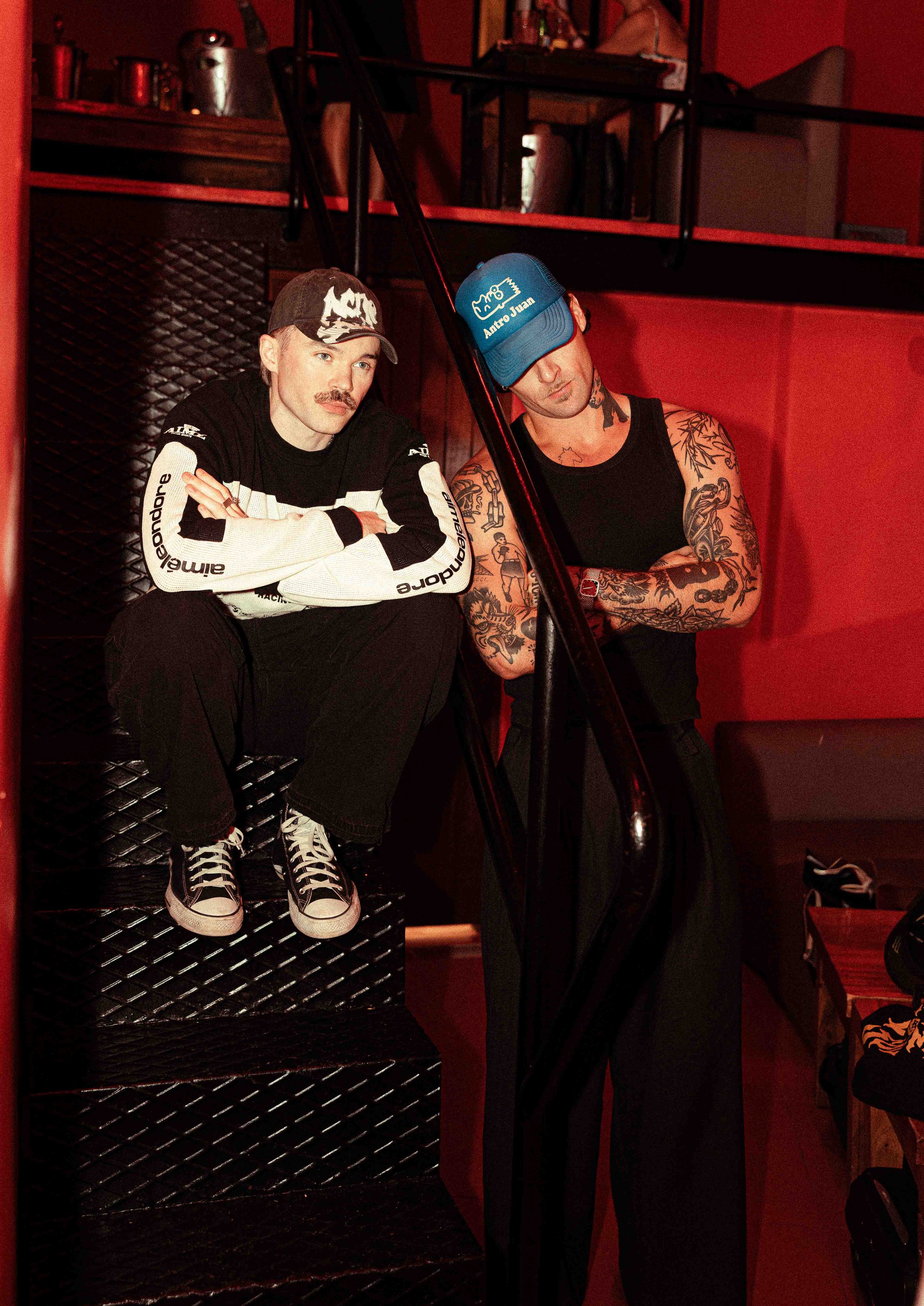 Two men sitting against a red wall; one with a mustache, wearing a black and white long sleeve shirt, a black cap, and sneakers, sitting on a black metal bench. The other man with tattoos on arms, wearing a blue cap, black sleeveless shirt, and black