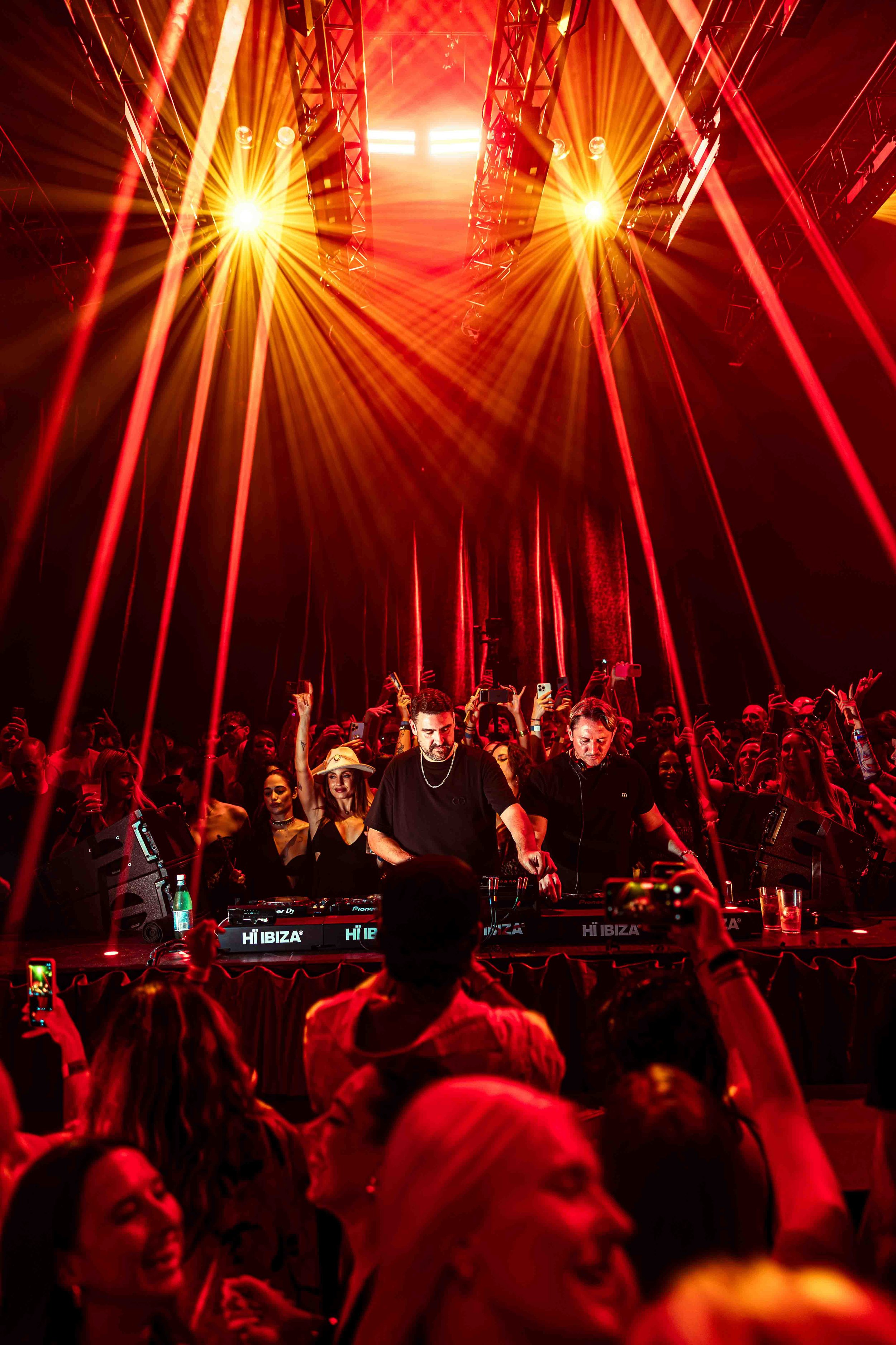 A DJ performing on stage at a nightclub with a large crowd of people dancing and taking photos in front. Bright red and yellow stage lights illuminate the scene.