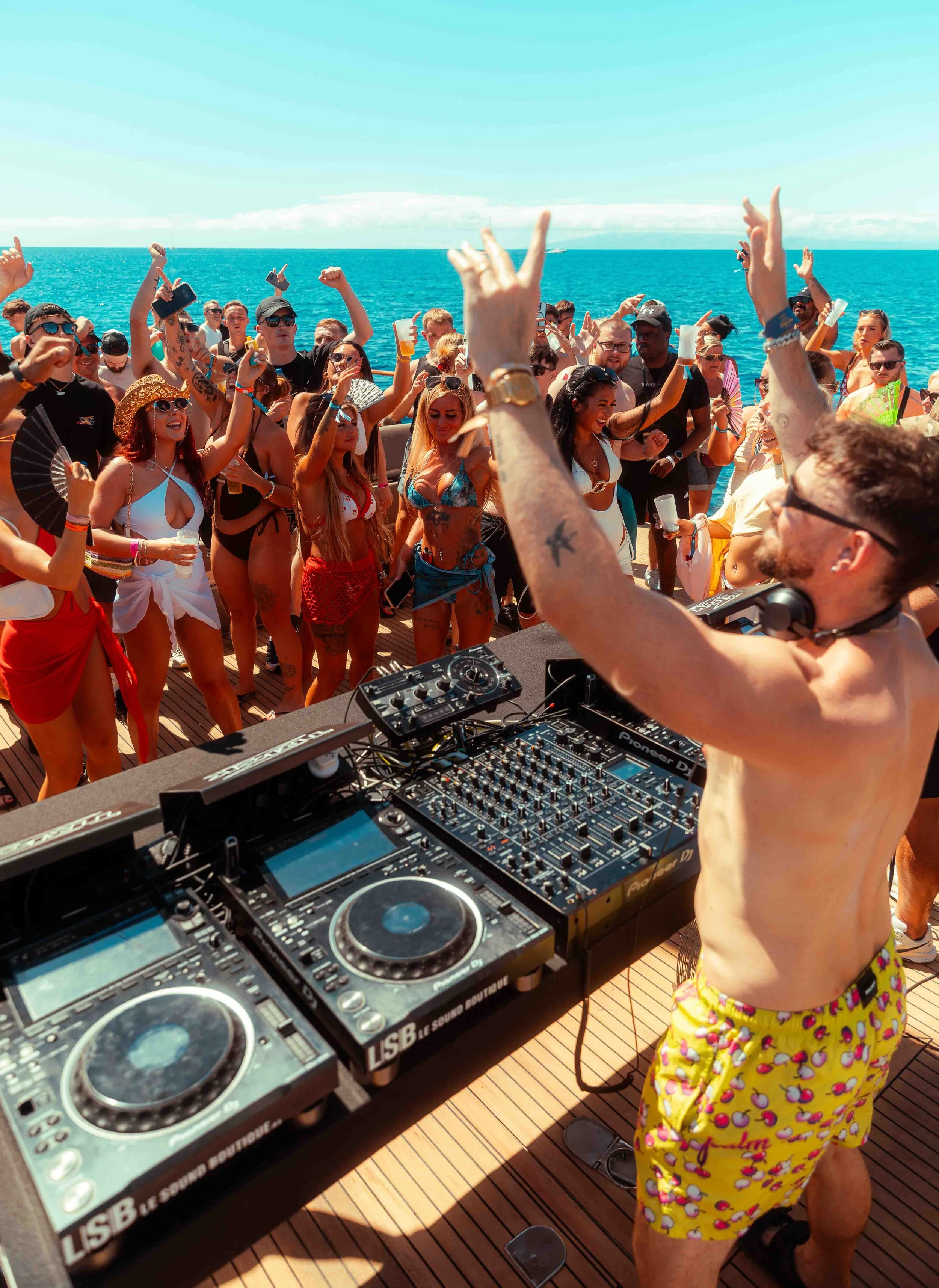 A diverse group of people dancing and enjoying music on a boat deck with the ocean and blue sky in the background. A DJ is playing music, and some people are holding drinks and celebrating.