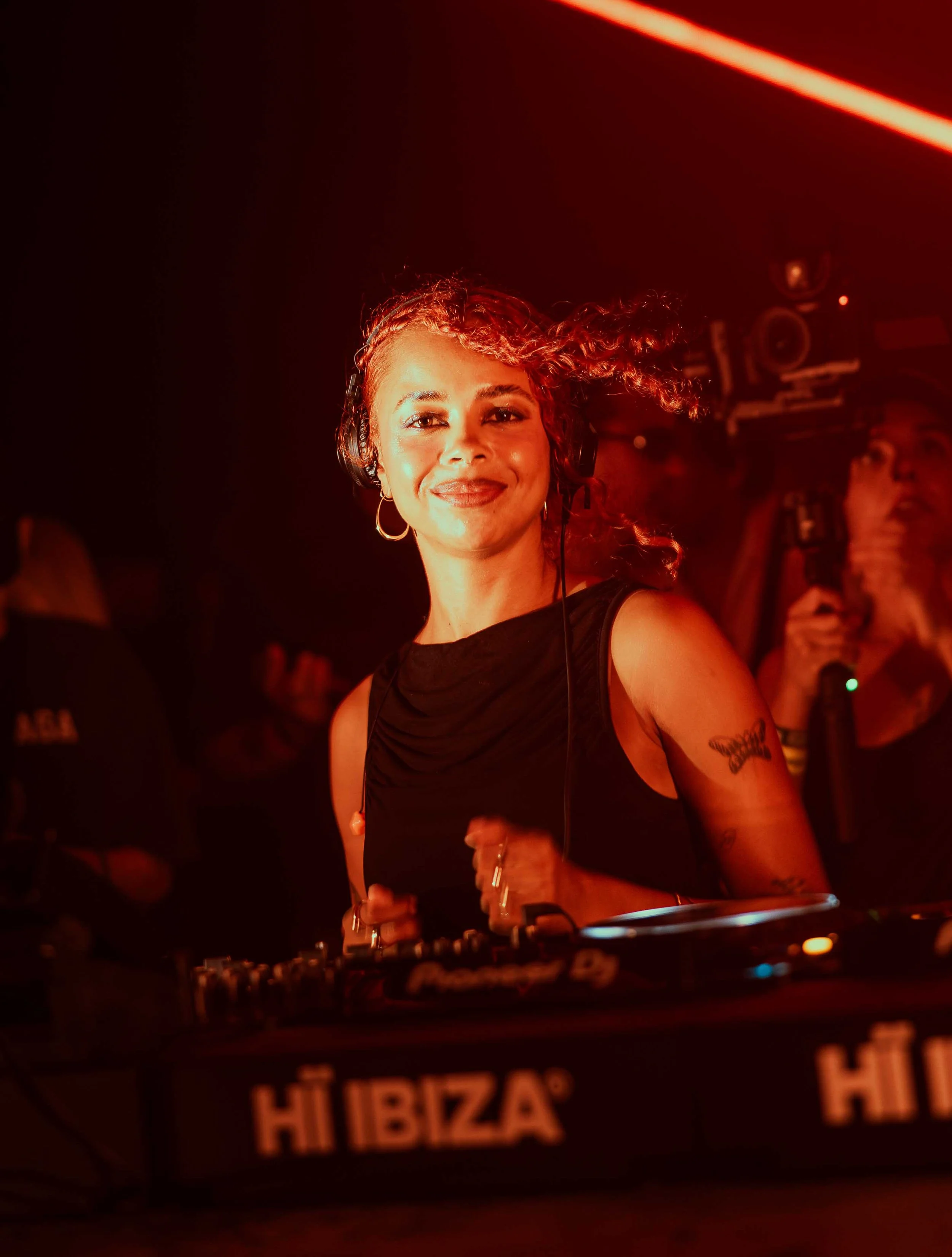 A woman with curly hair and earrings smiling and DJing at a nightclub with red lighting and a behind-the-scenes view of DJ equipment.