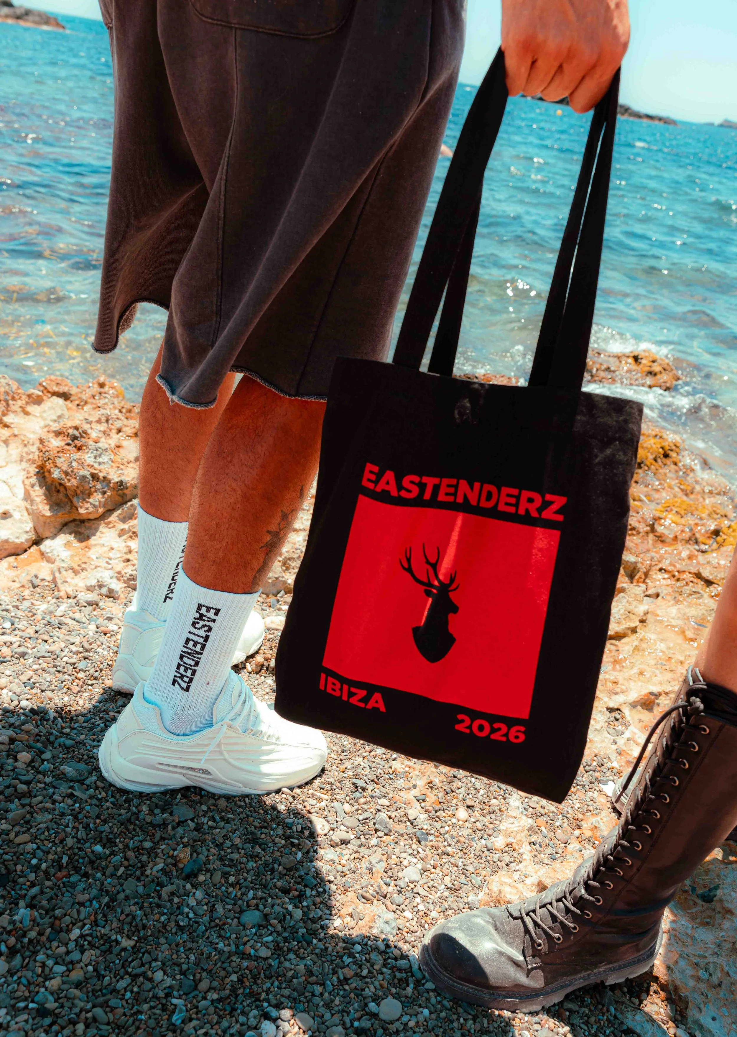 Close-up of a person's legs and feet standing on a rocky beach near the water. The person is wearing white sneakers and white socks that say 'EASTENDERZ'. They are holding a black tote bag with red and black artwork and text that reads 'EASTENDERZ IB