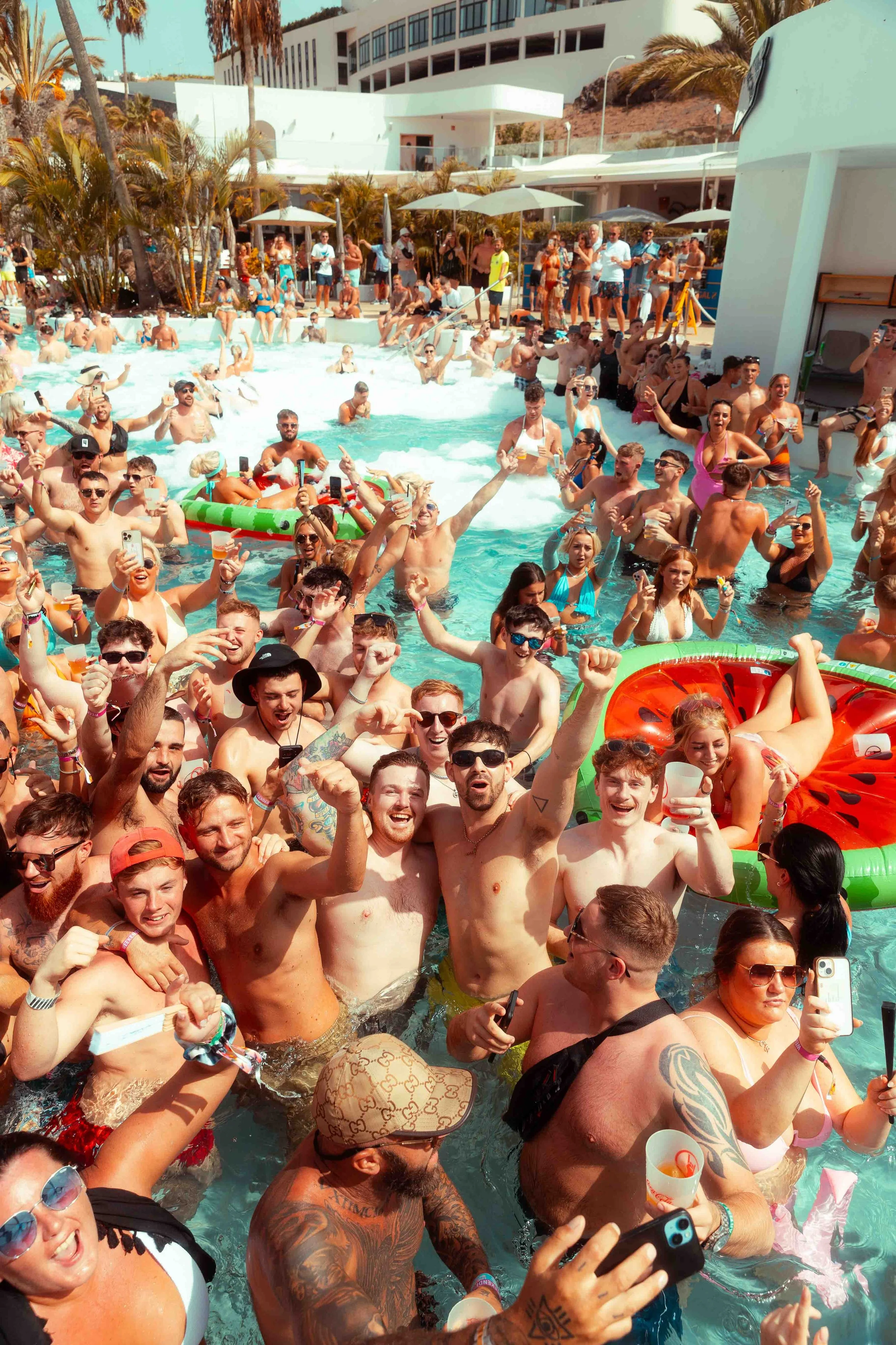 People gathered in a pool, celebrating and taking photos, with some holding drinks. The scene is lively with many smiling faces, surrounded by palm trees and a modern building in the background on a sunny day.