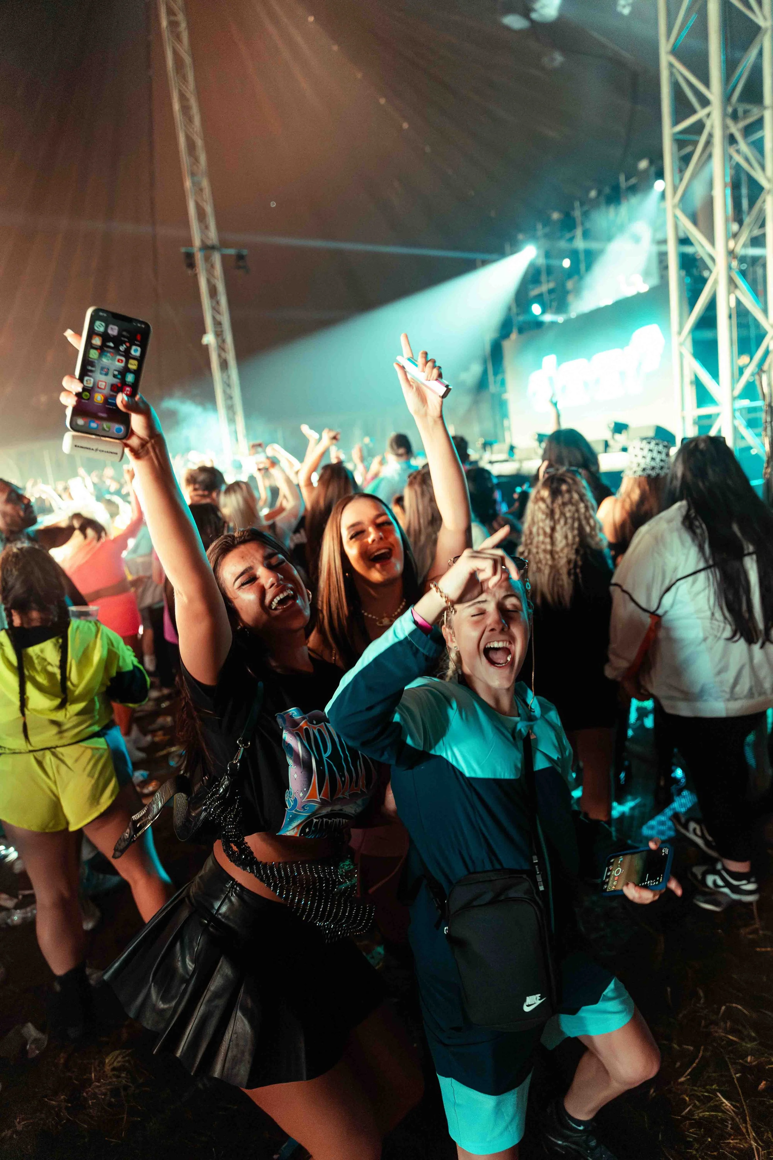A group of people at a concert or music festival, dancing and enjoying themselves under stage lights with a large screen in the background.
