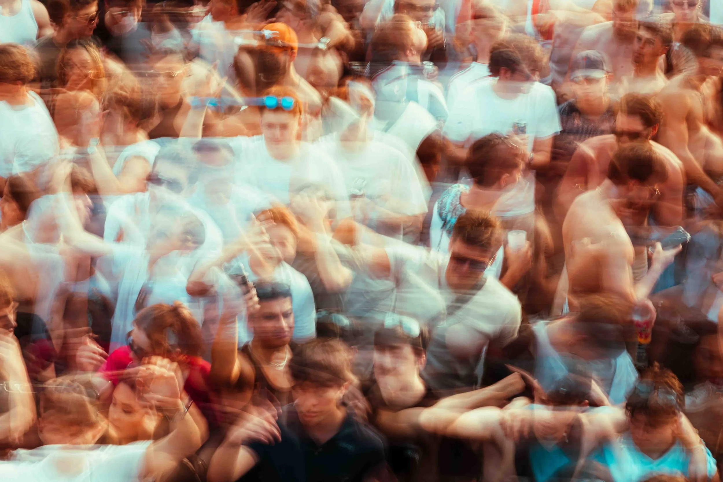 A large crowd of people at an outdoor event with some people wearing sunglasses and holding drinks, captured with motion blur.