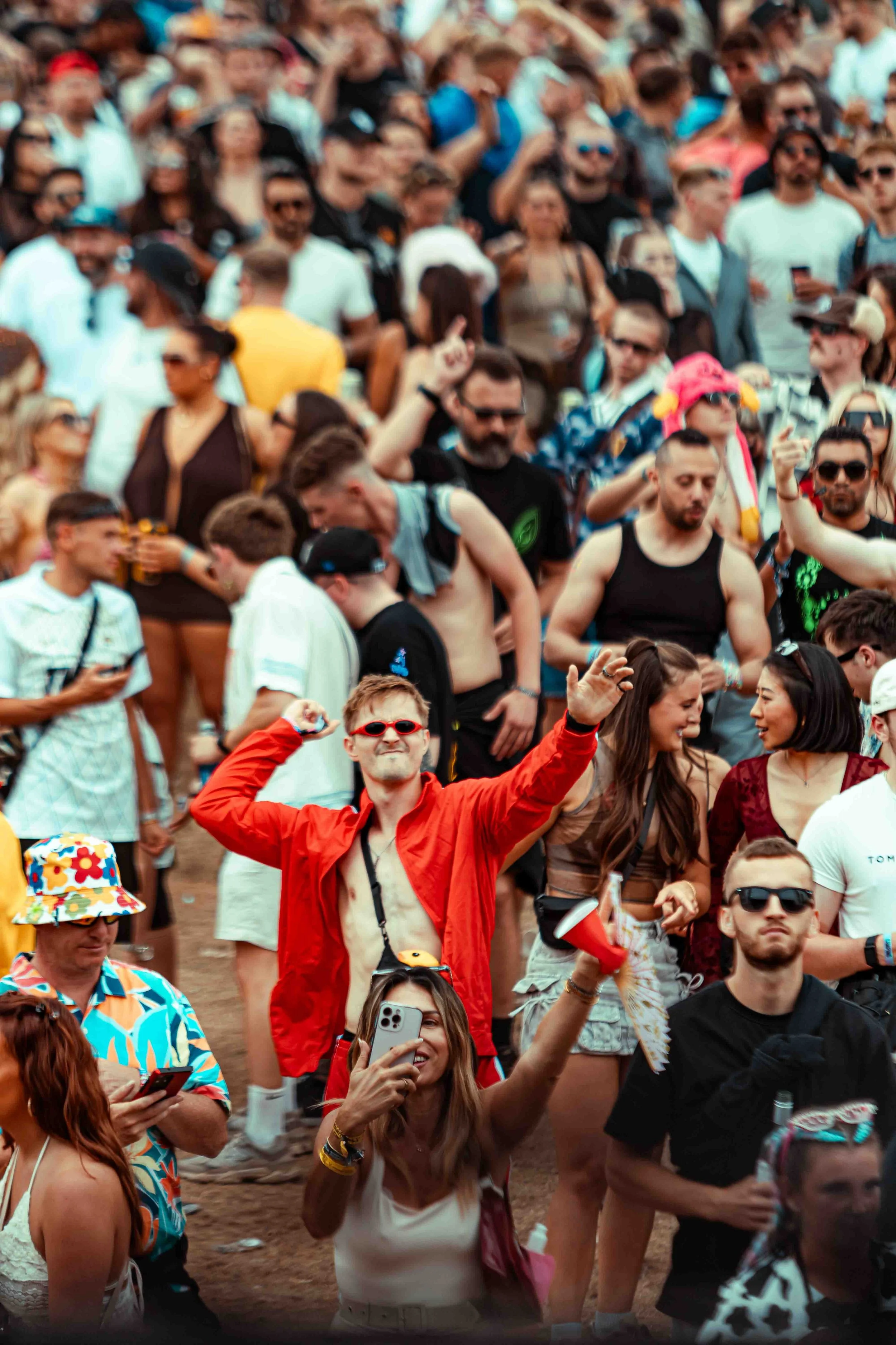 A large crowd of people attending an outdoor event, many wearing sunglasses and summer clothing, some holding phones, and one person in the foreground wearing a red jacket and sunglasses.