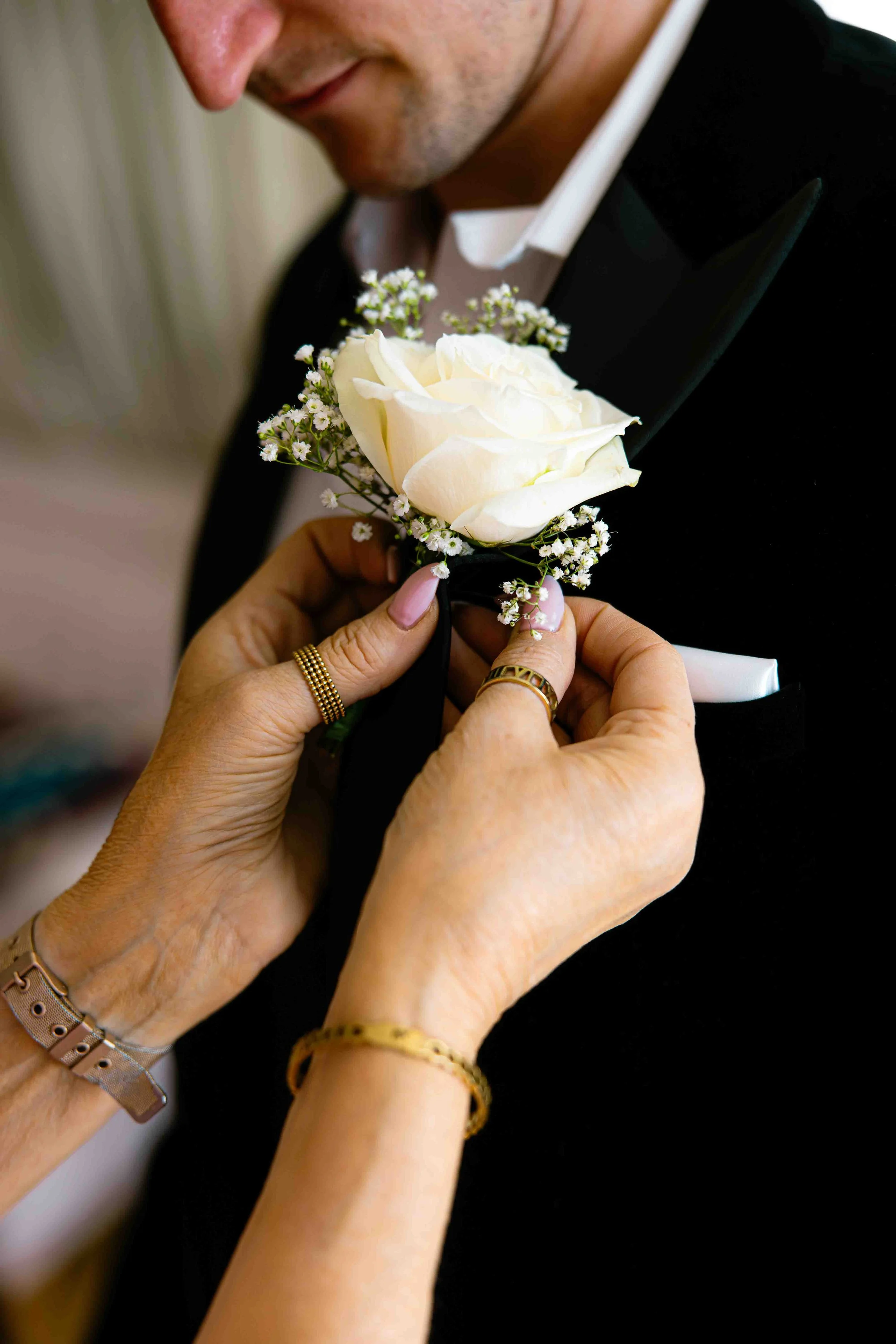 Close-up of a man in a tuxedo with a white shirt, receiving a white rose and baby's breath boutonniere pinned to his lapel from two women's hands, one with gold rings and the other with a rose gold watch and chain bracelet.