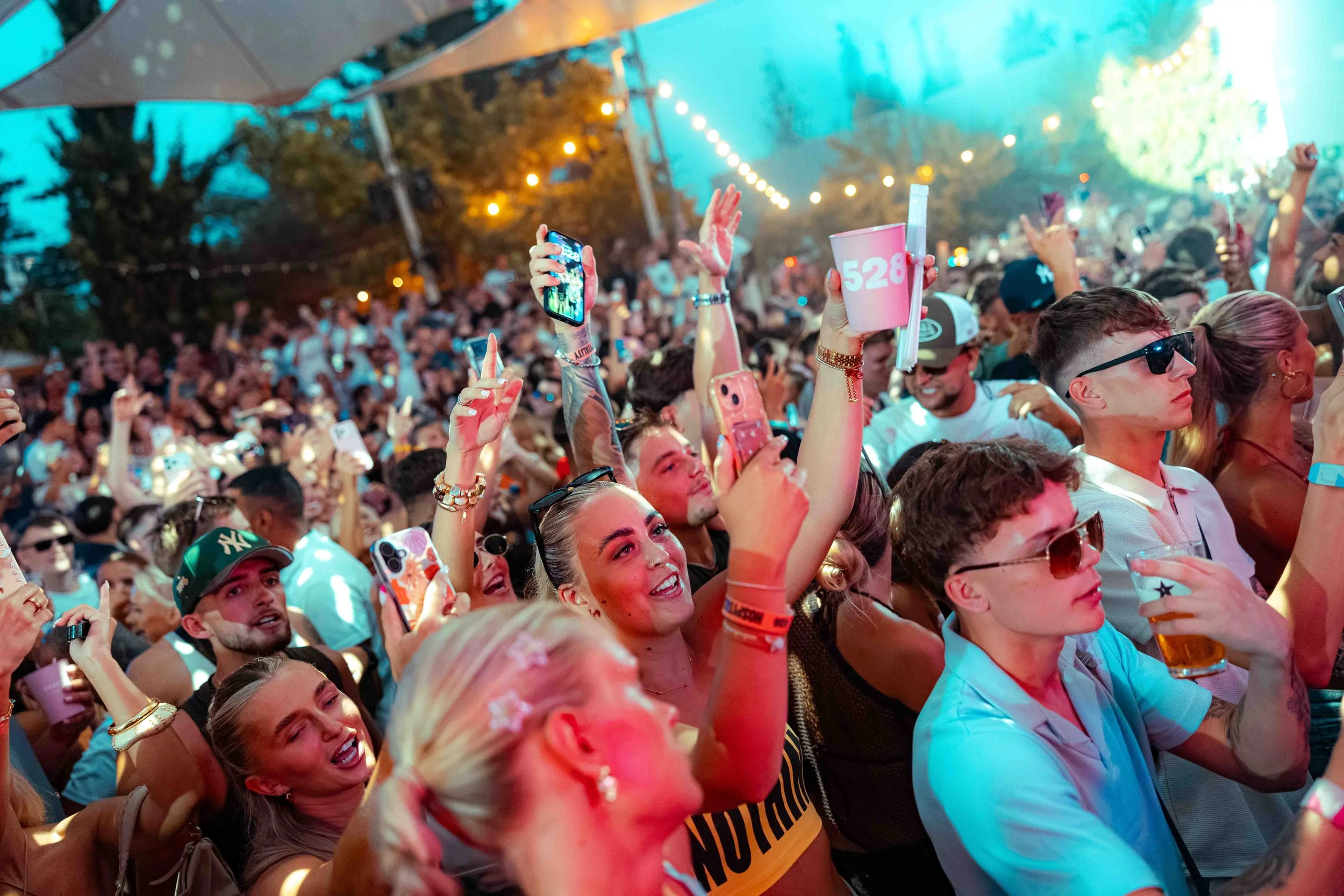 Crowd of people at an outdoor event with many raising their hands and holding phones, with colorful lighting and string lights overhead.