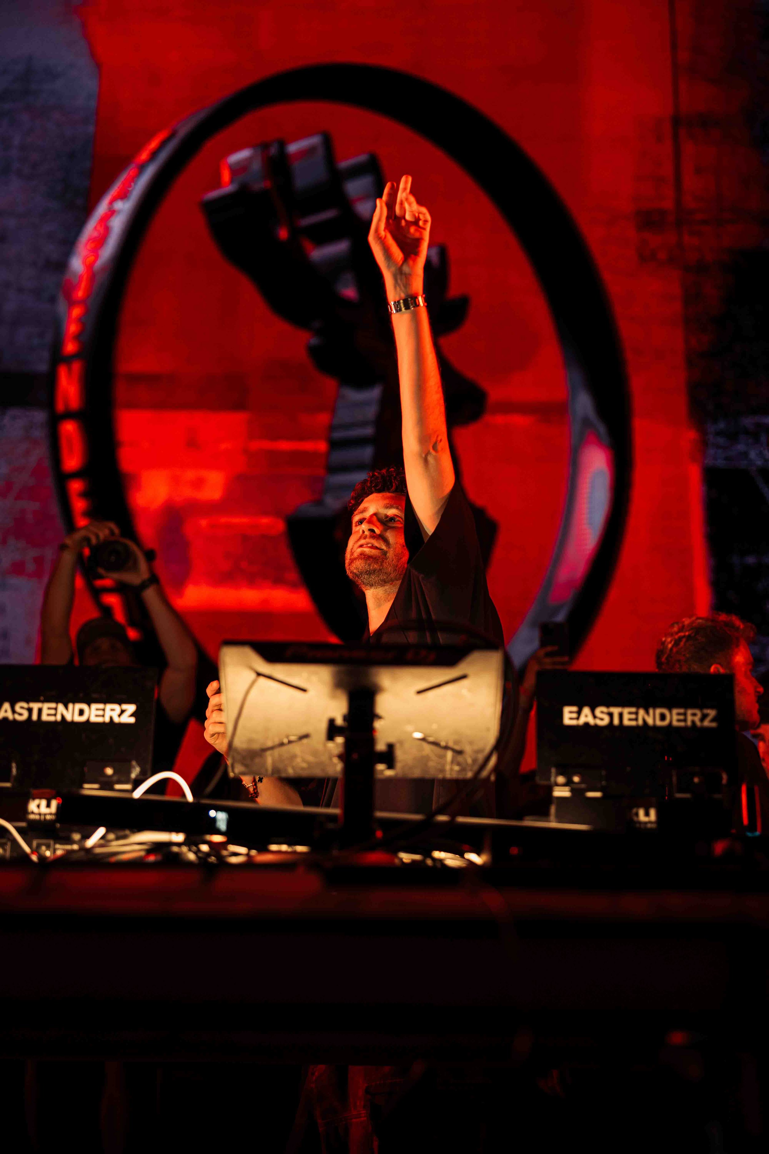 A DJ performing at a concert with a large sculpture of a raised fist behind him, illuminated with red lighting, and DJ equipment labeled 'EASTENDERZ' in front.