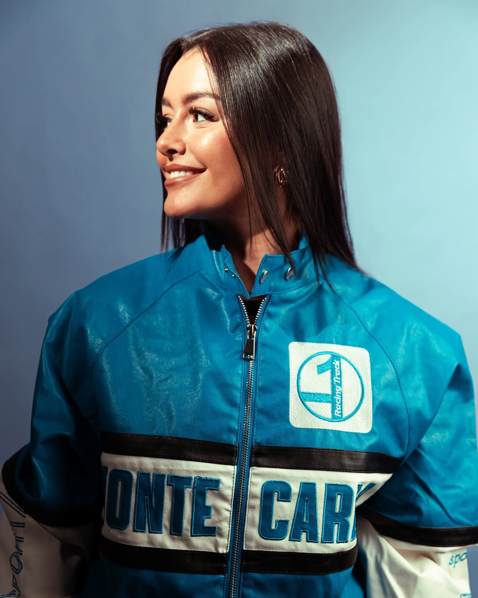 Close-up of a smiling woman with shoulder-length dark brown hair, wearing a blue and black jacket with large embroidered text.