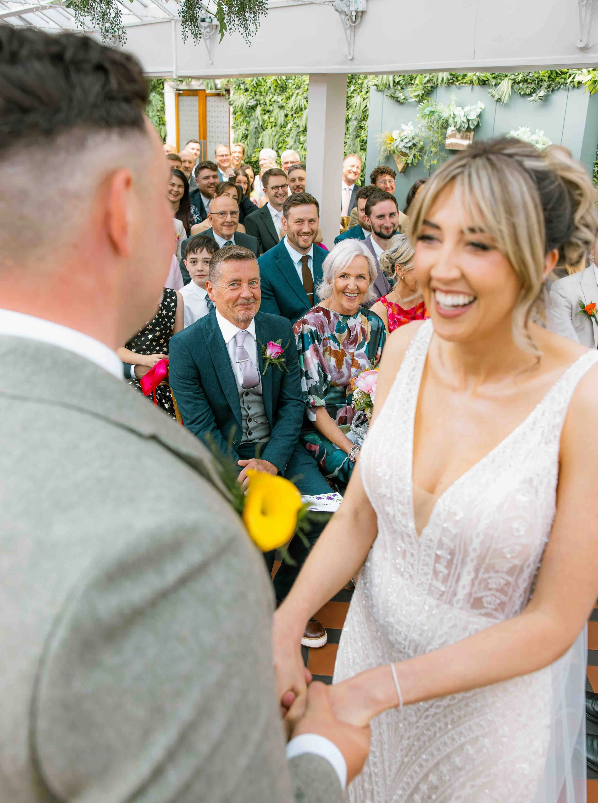 A bride and groom holding hands during their wedding ceremony with guests looking on and smiling in a lush, green indoor venue.