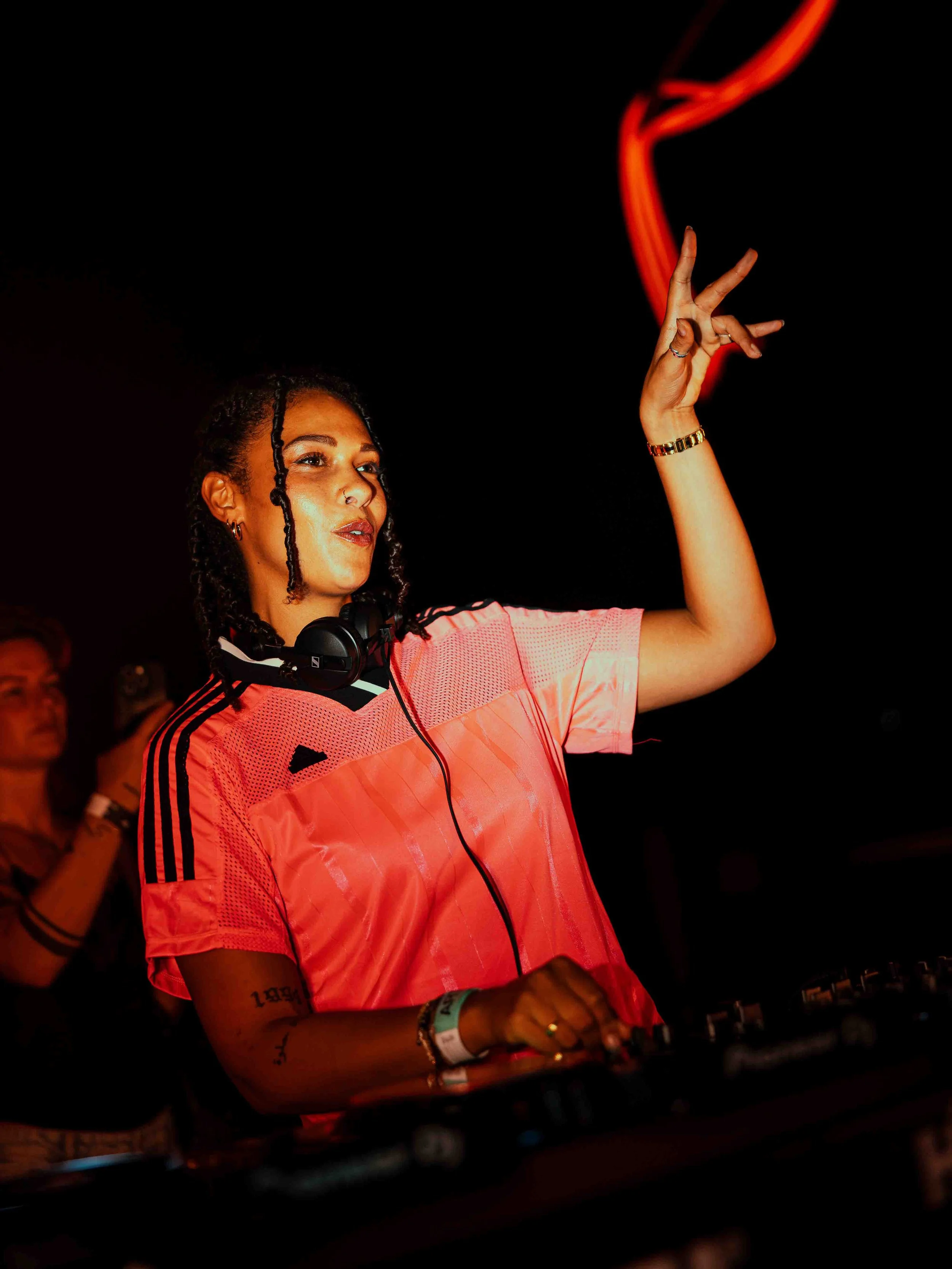 A woman with dreadlocks wearing a pink Adidas shirt, headphones around her neck, and a gold watch, is DJing at a dark indoor event.