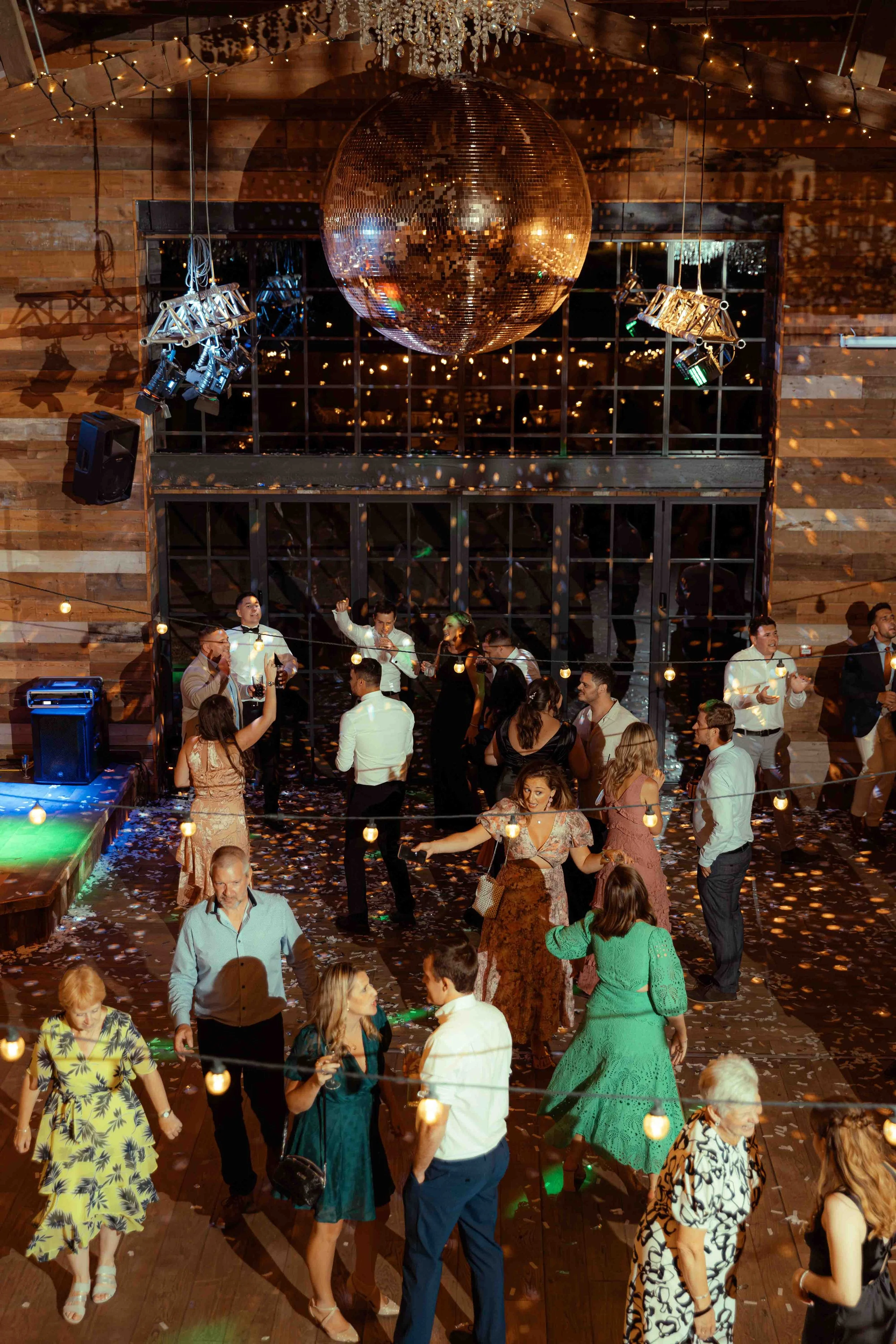 People dancing and socializing at a party with a large gold disco ball, string lights, and confetti on the floor. The setting has a rustic, wooden interior.