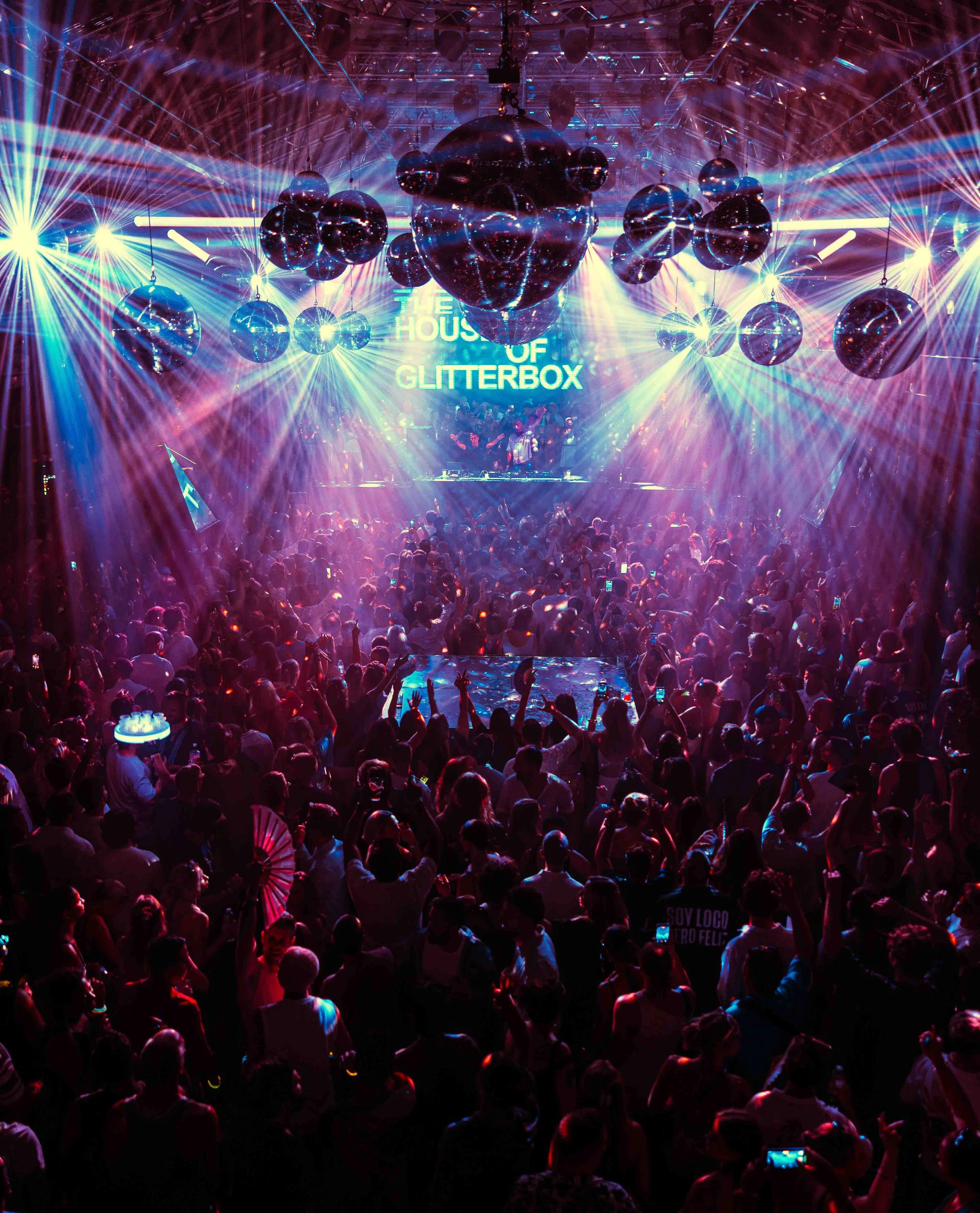A crowded dance floor at a nightclub with colorful laser lights and multiple large disco balls hanging from the ceiling, illuminated by bright stage lights, and a DJ playing music behind a booth with a sign that reads 'The House of Glitterbox'.