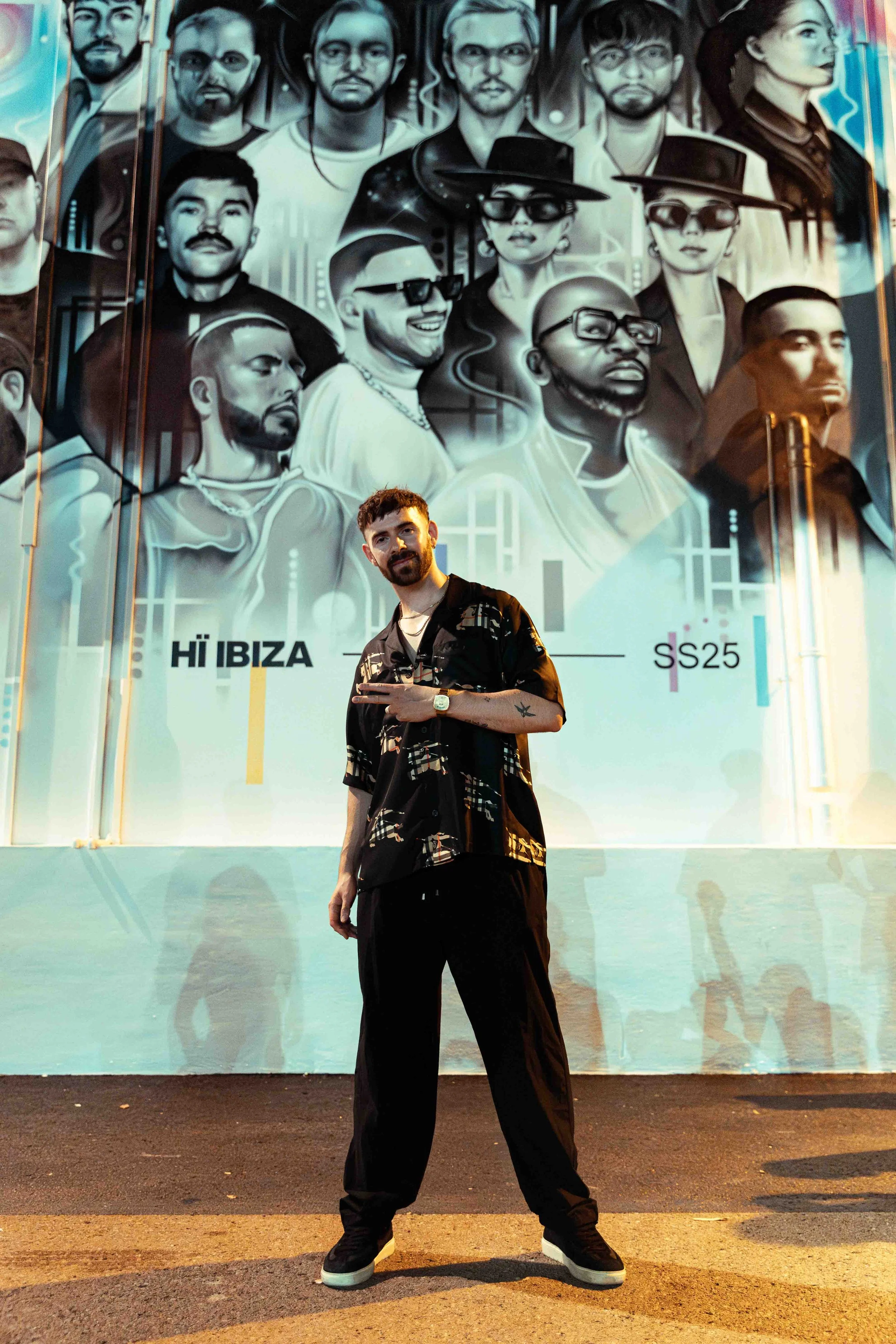 A man with a beard and short hair standing in front of a large grayscale mural of diverse people's faces, featuring stylish clothing and accessories, in an outdoor urban setting at night.