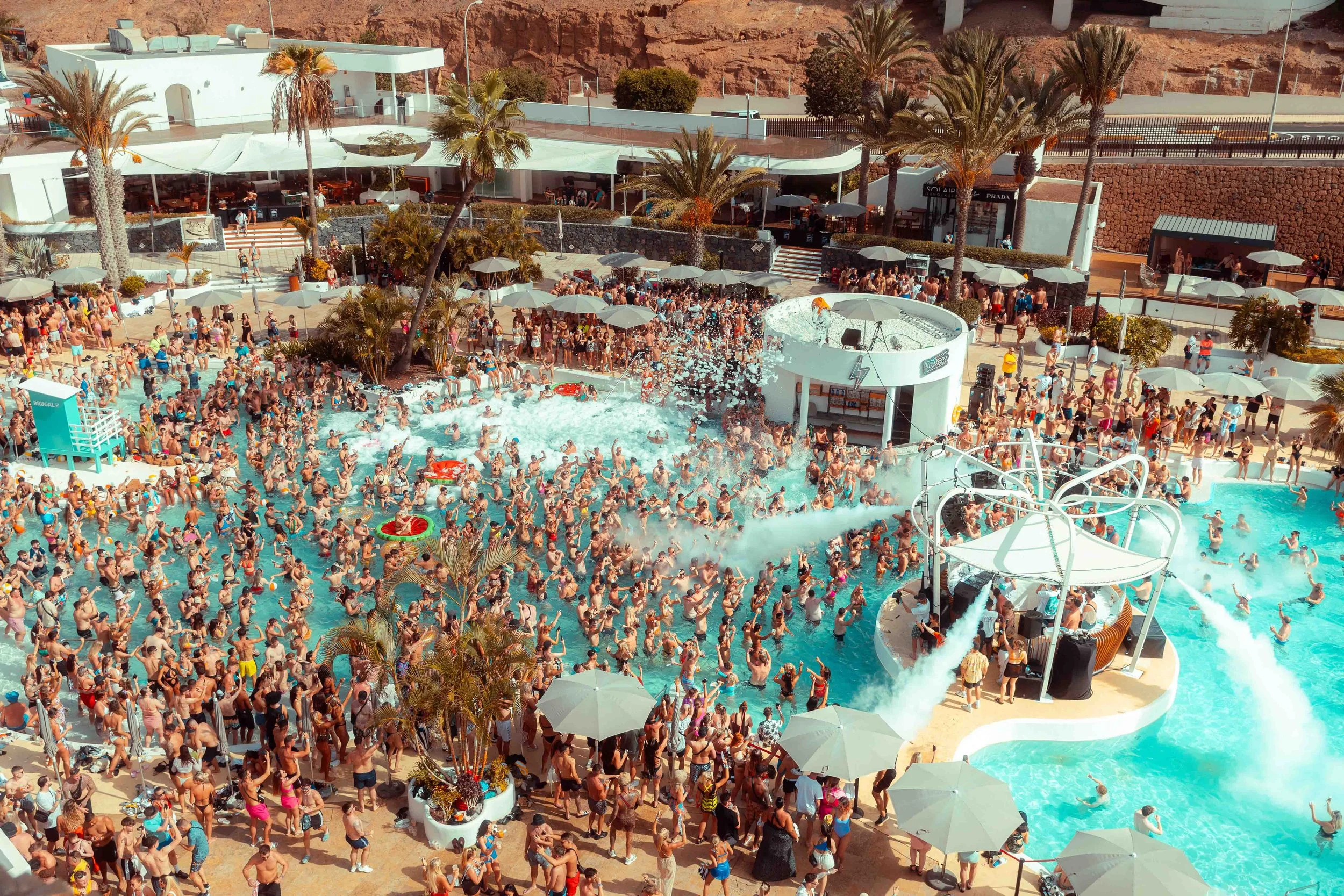 A crowded outdoor swimming pool party with many people enjoying the pool, some on the slides, and others lounging under umbrellas, surrounded by palm trees and a modern building with white architecture.