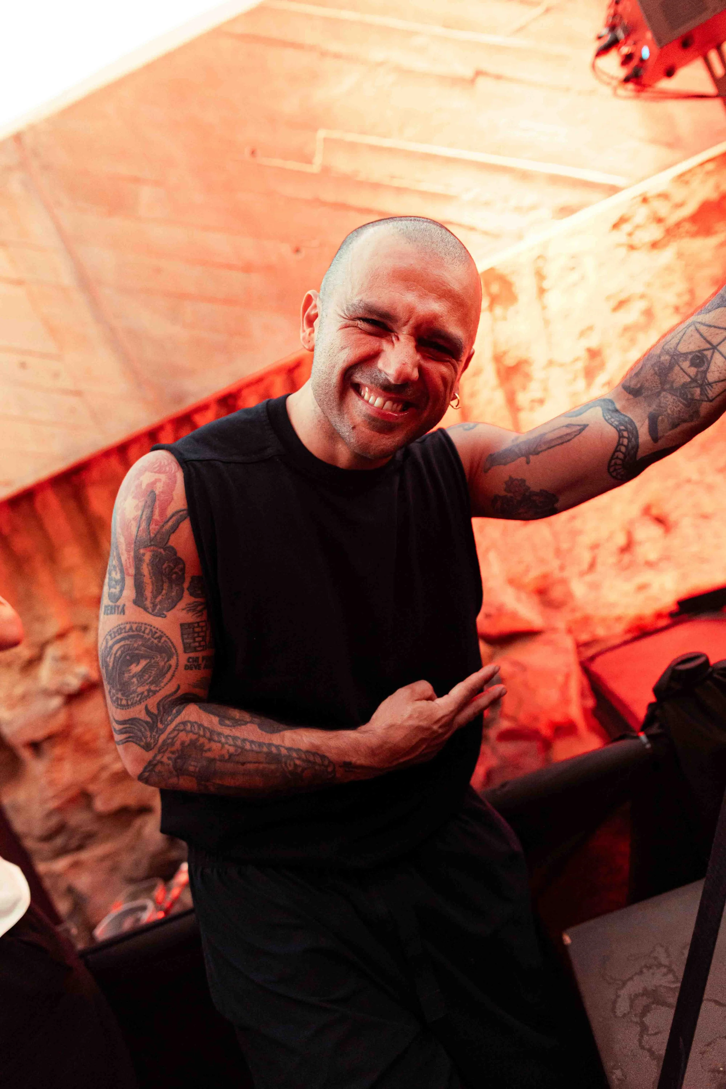 A man with a shaved head and multiple tattoos on his arms, wearing a black sleeveless shirt, is smiling broadly and squinting his eyes while making a peace sign with his right hand. He is inside a room with wooden walls and is in front of some audio 
