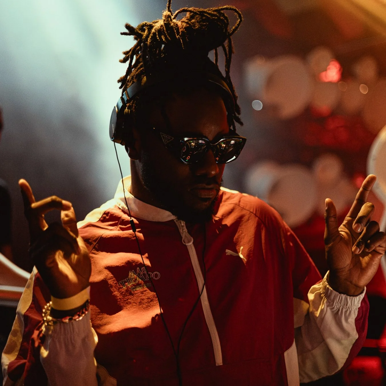 A man with dreadlocks wearing sunglasses, a red jacket, and headphones, dancing or enjoying music in a dimly lit environment.