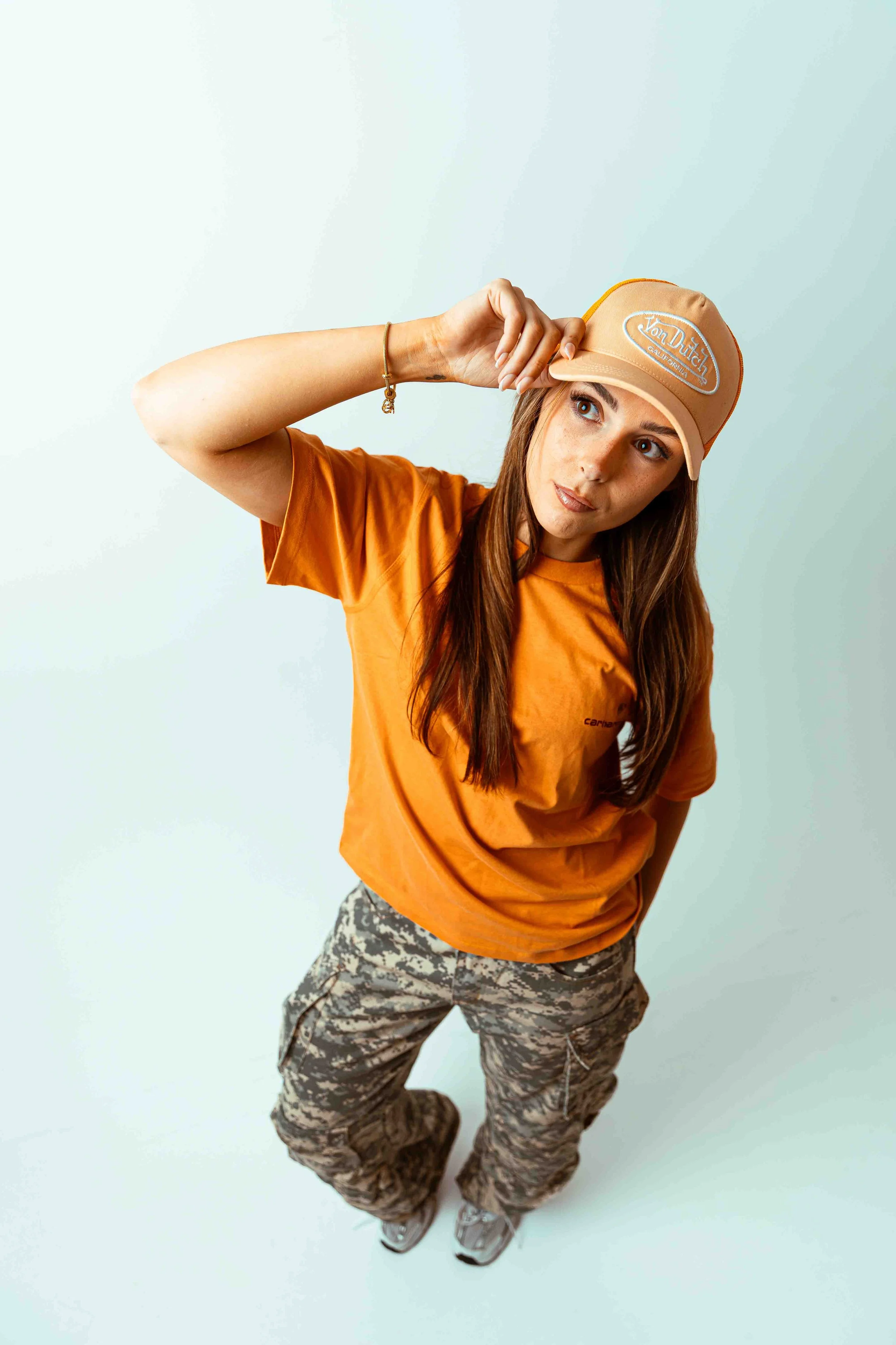 Emma 2000 with brown hair wearing a beige 'Von Dutch California' baseball cap, orange T-shirt, camouflage cargo pants, and sneakers, standing against a plain light-colored background.