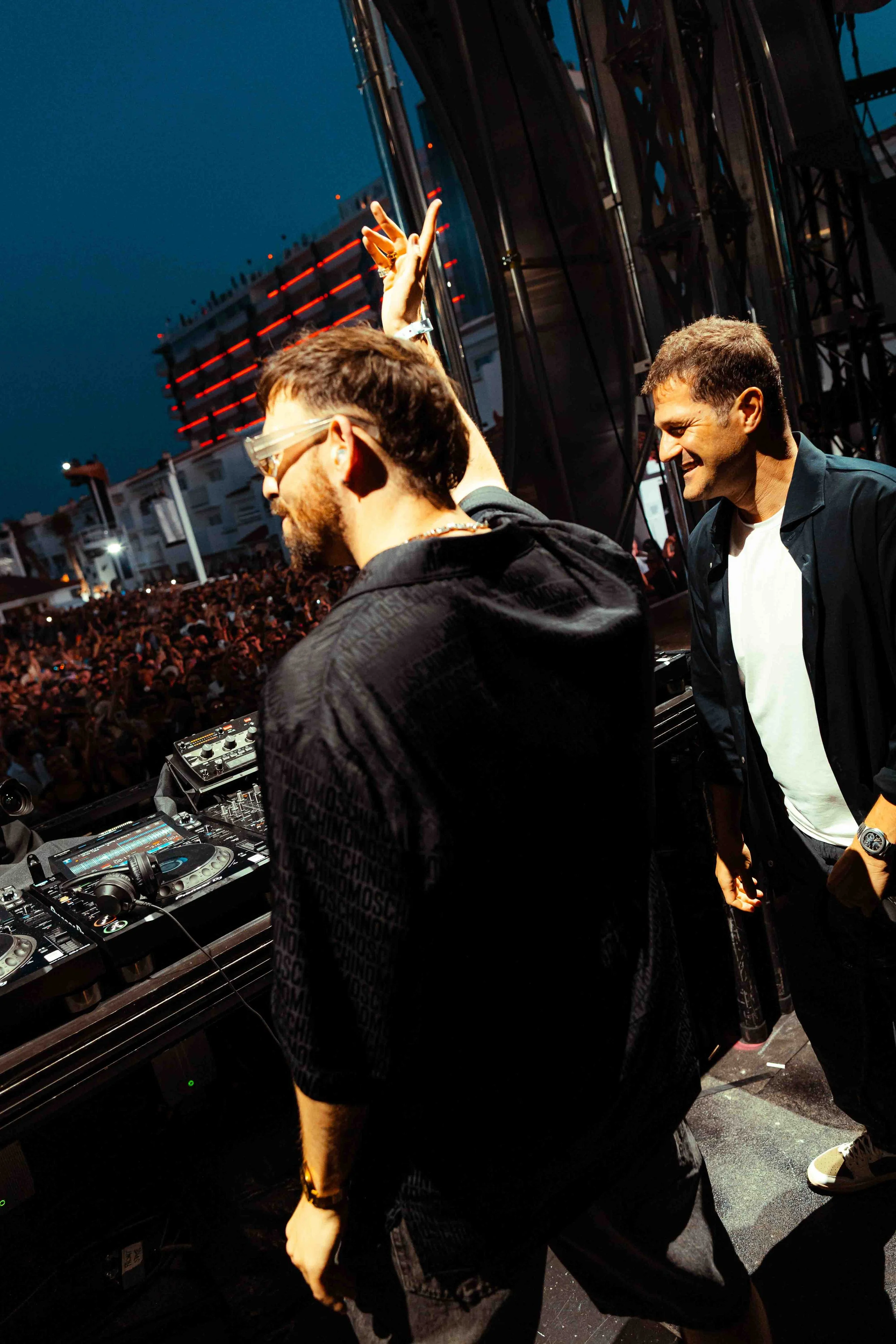 A DJ wearing glasses and a black jacket performs at an outdoor event at night, with a large crowd in the background. He is focused on the mixing console, while another man beside him smiles and watches.