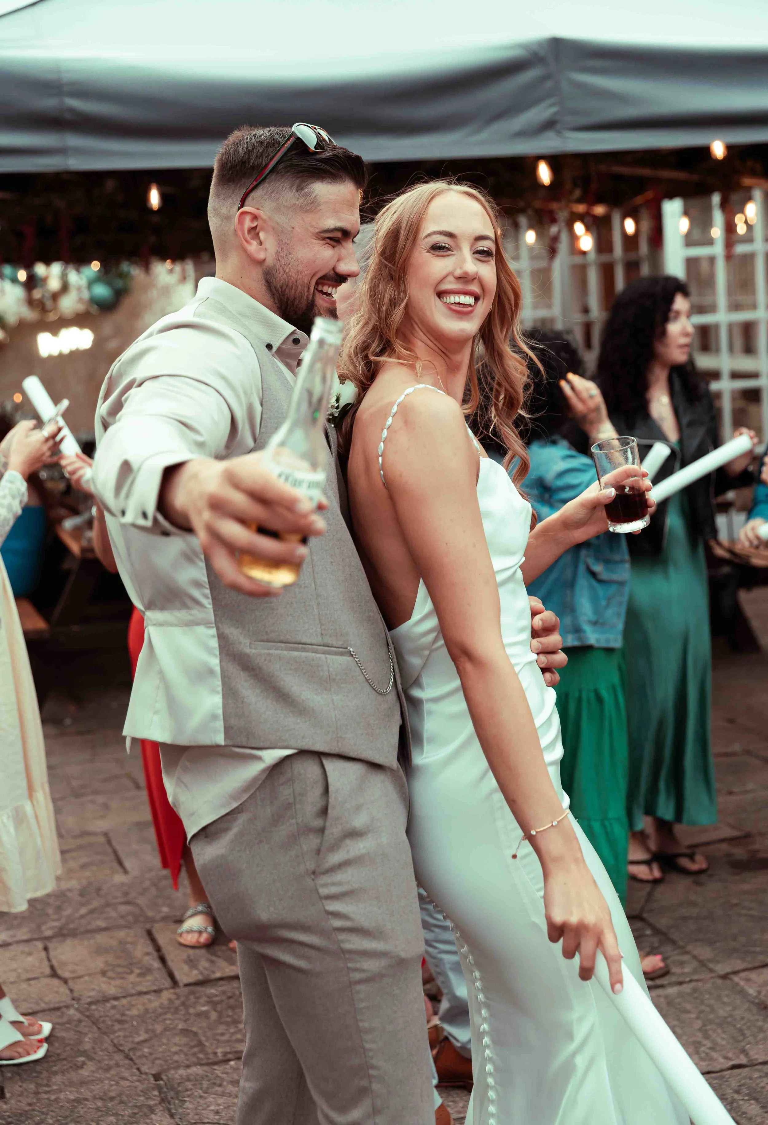 A smiling bride and groom at their wedding reception, holding drinks and dancing outdoors under a tent, with guests in the background.