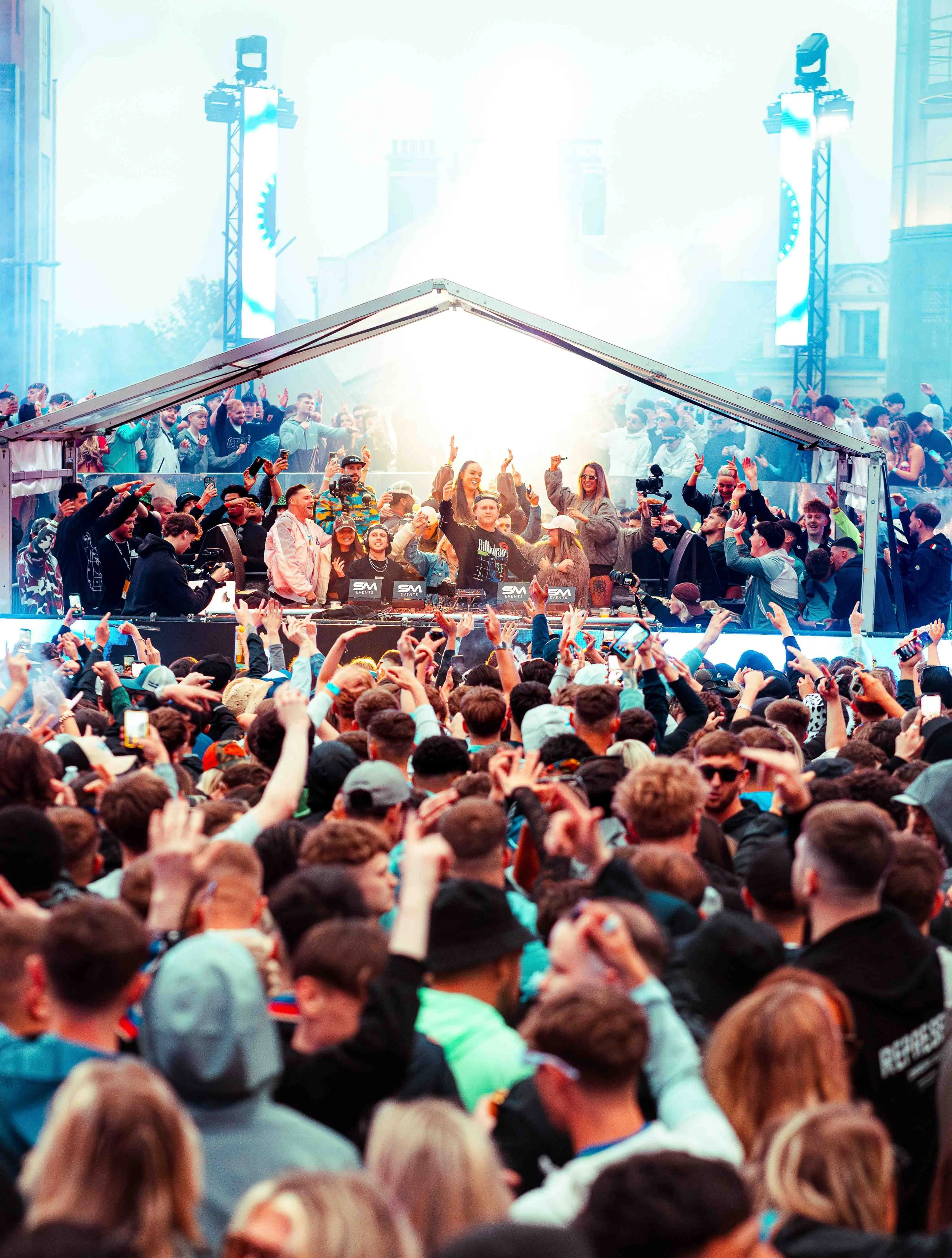 Max and Luke dean play to a crowd of people at a music or festival event with a DJ or performer on stage, with large speakers, lighting, and cheering attendees.