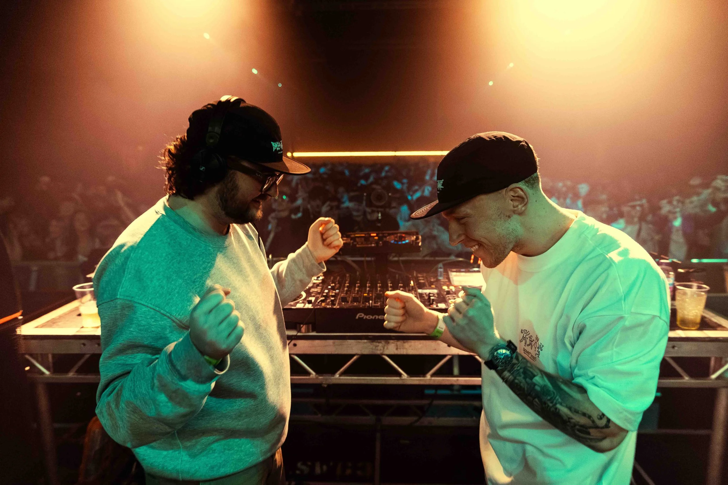 Two men in casual clothing and black hats, wearing headphones and wristbands, are on a dance floor facing each other with clenched fists, smiling and appearing excited. There is a DJ setup with turntables behind them and the background is filled with