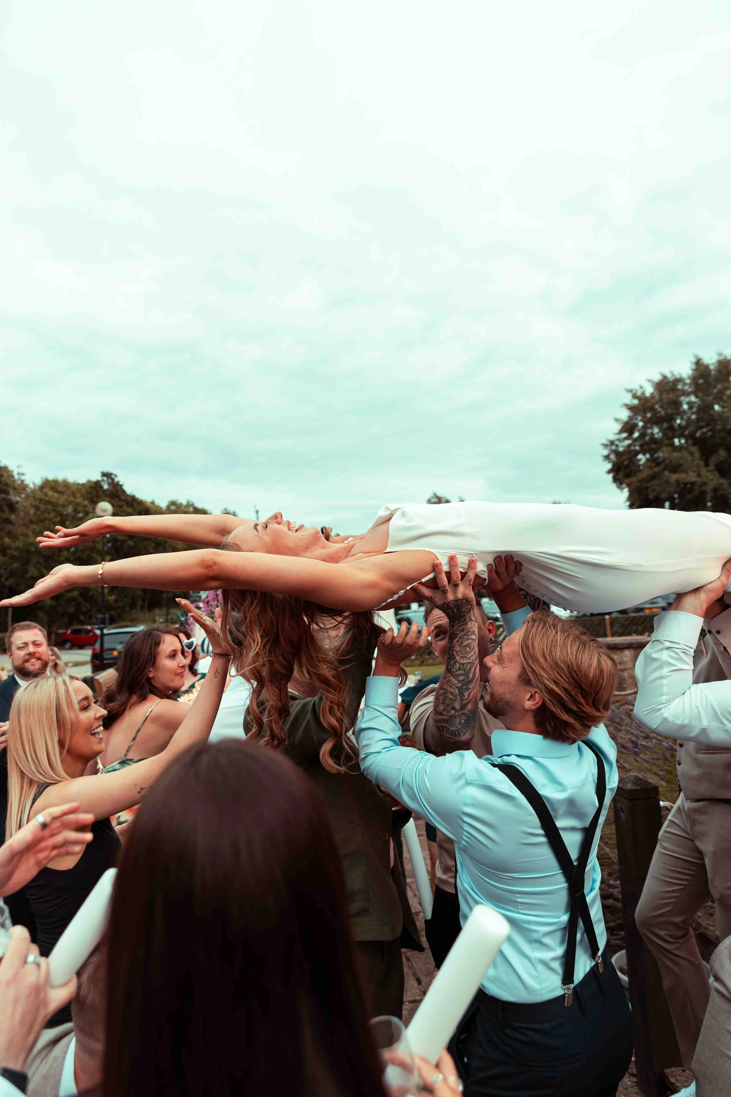 Group of people celebrating outdoors as a woman in a wedding dress is being lifted by others during a wedding celebration.