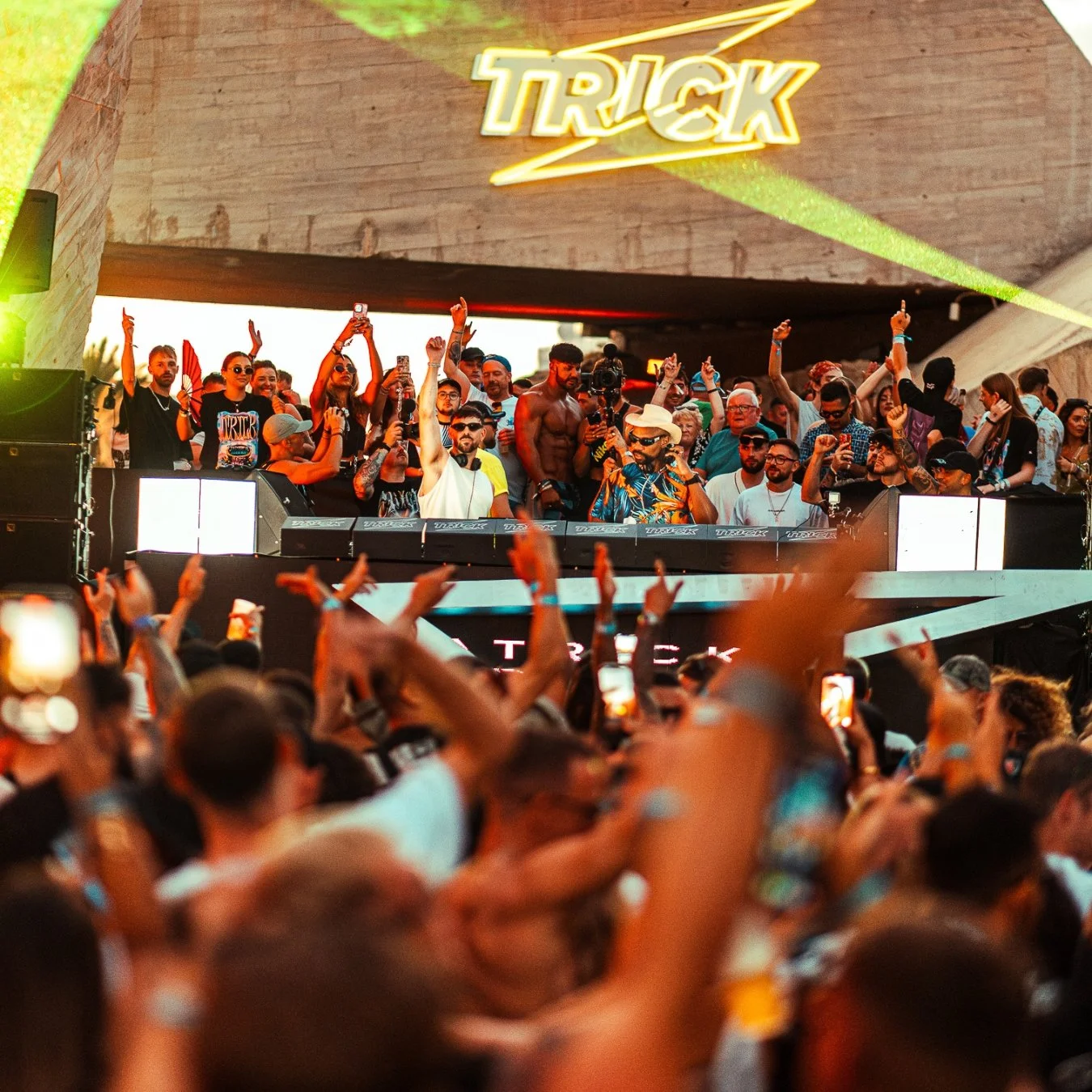 Crowd of people at an outdoor music festival or concert with a DJ on stage, some raising their hands and taking photos or videos, with a large 