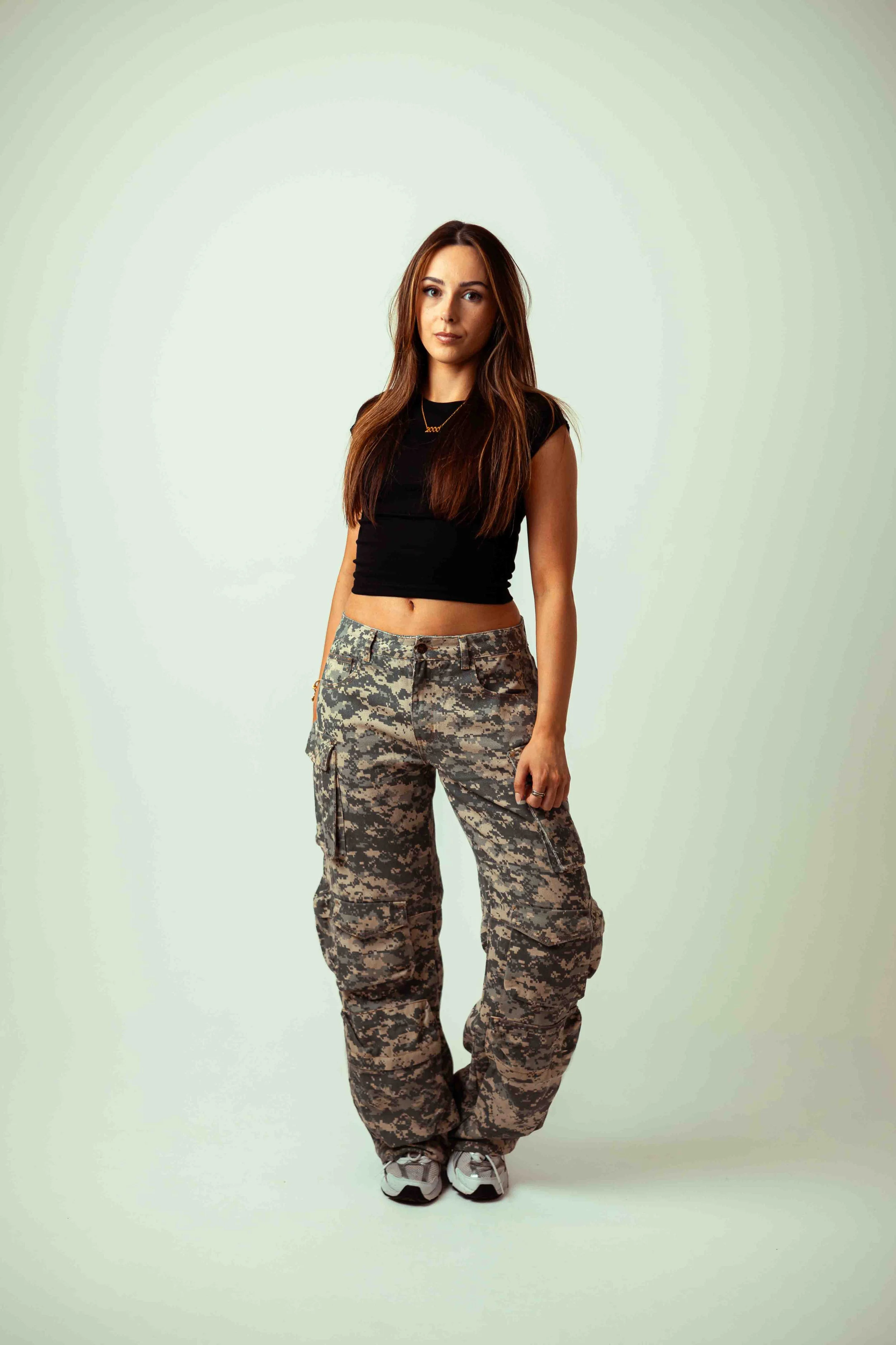 Emma 2000 standing in front of a plain light-colored background, wearing camouflage cargo pants, a black crop top, and white sneakers, with long brown hair and a confident expression.