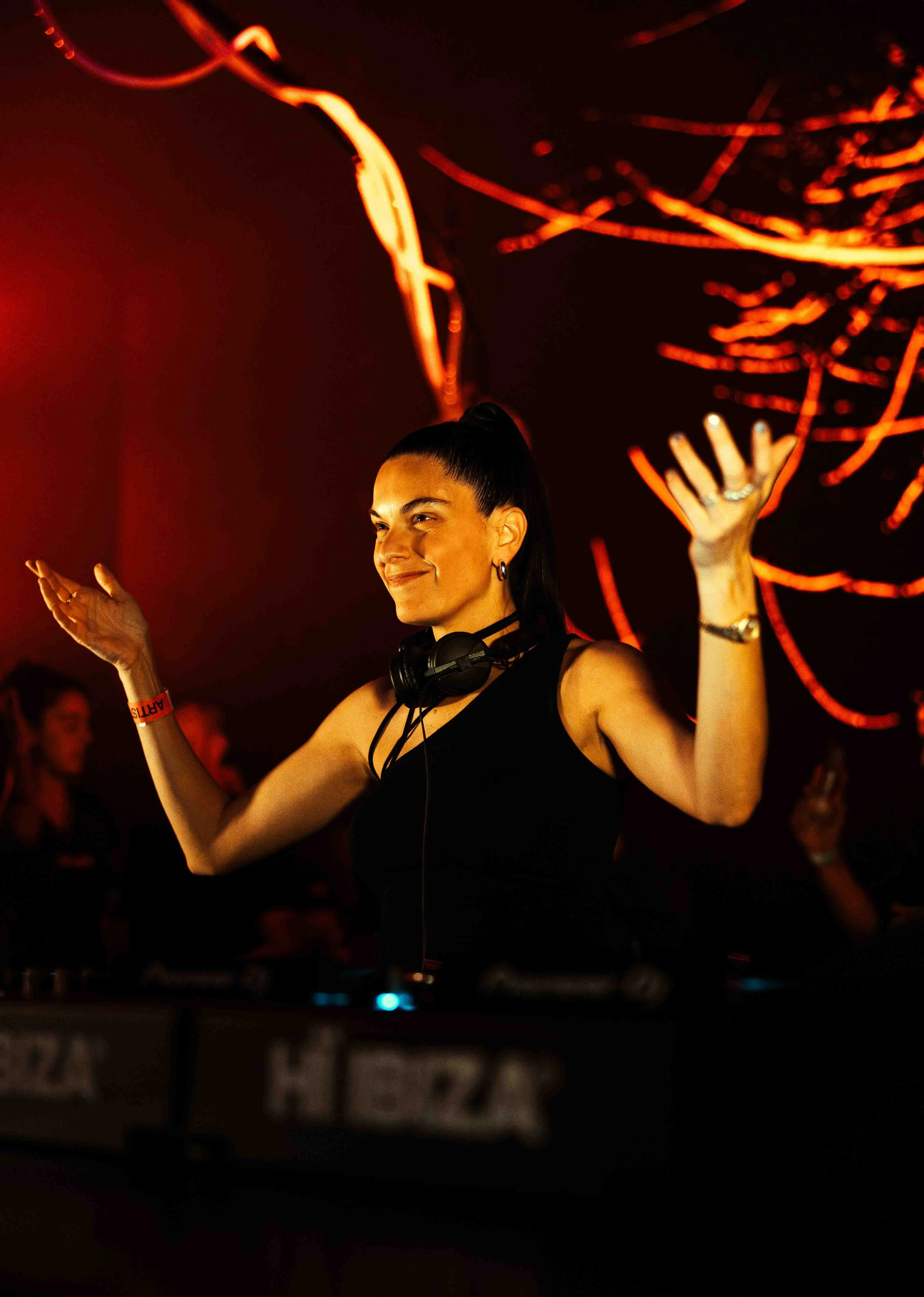 A female DJ with dark hair tied back in a ponytail, wearing headphones around her neck, dancing with a smile and her hands raised in the air at a nightclub or music event with red lighting and blurred figures in the background.