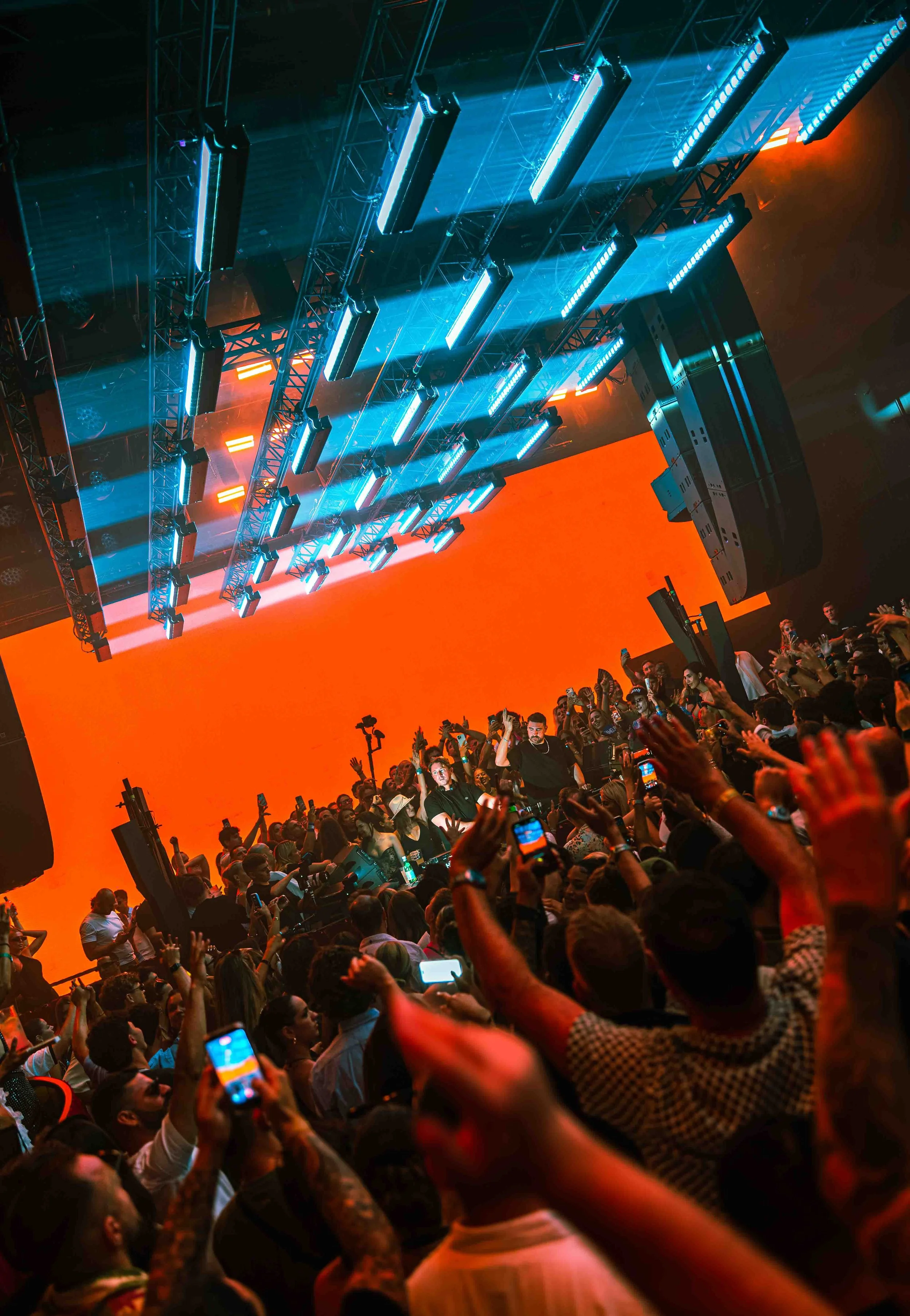 A large crowd of people at a concert or event, with many holding up their smartphones, on a stage with bright blue lights and an orange background, Camelphat playing at HI ibixa