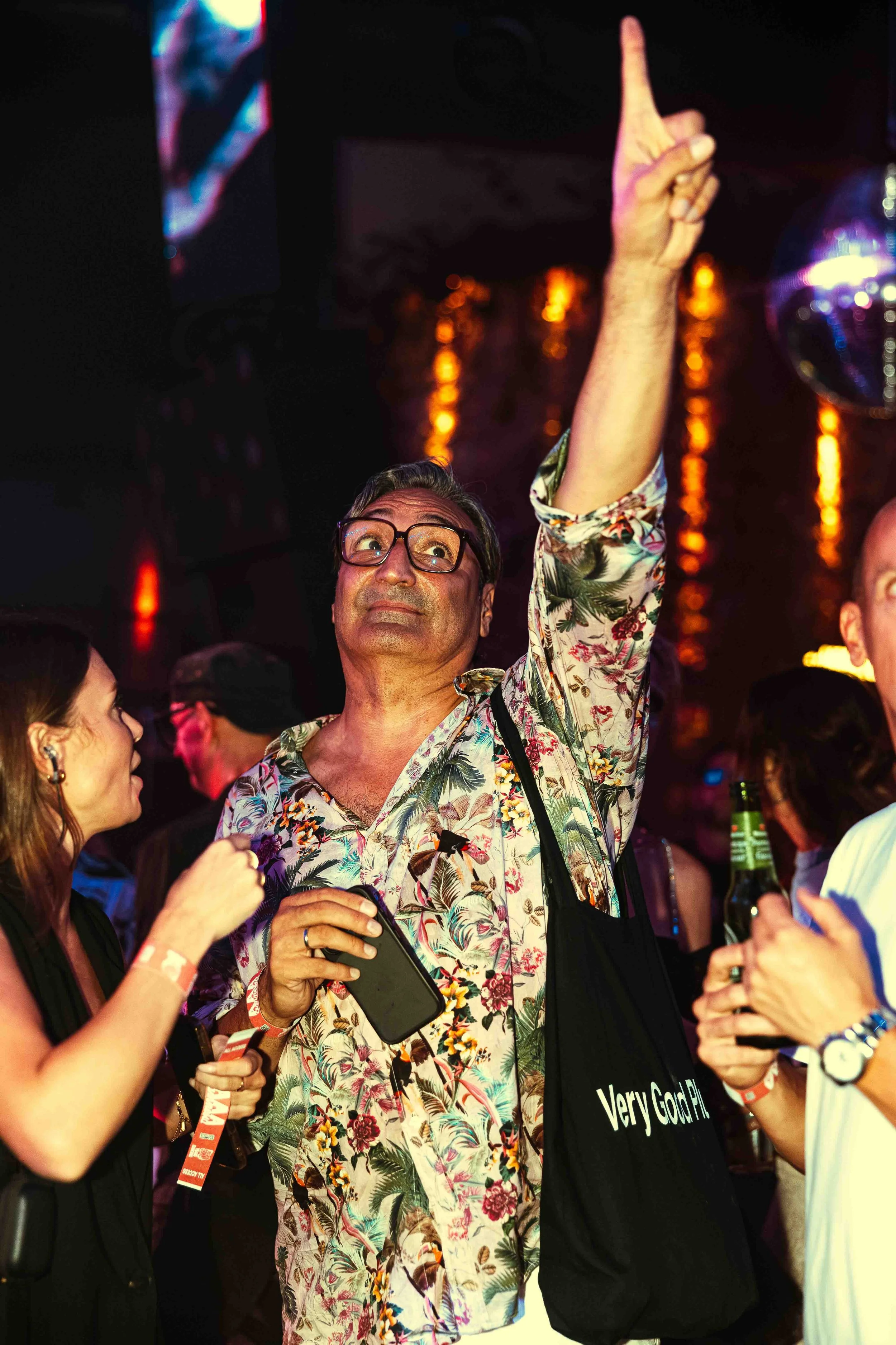 A man wearing a floral shirt and glasses raising his right hand to point upward, surrounded by people, in a dimly lit environment with colorful reflections.