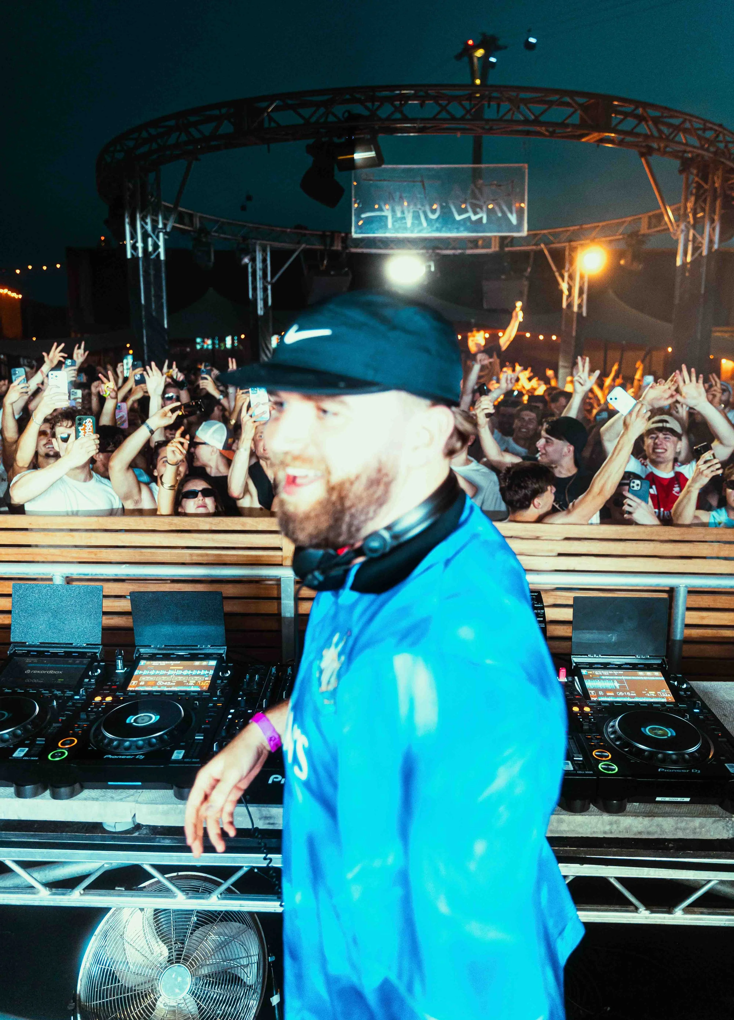 Gaskin DJ wearing a blue shirt and black Nike hat performing at a crowded outdoor music event during evening or night, with a large audience raising their hands and taking photos, under a stage with lighting and a neon sign.