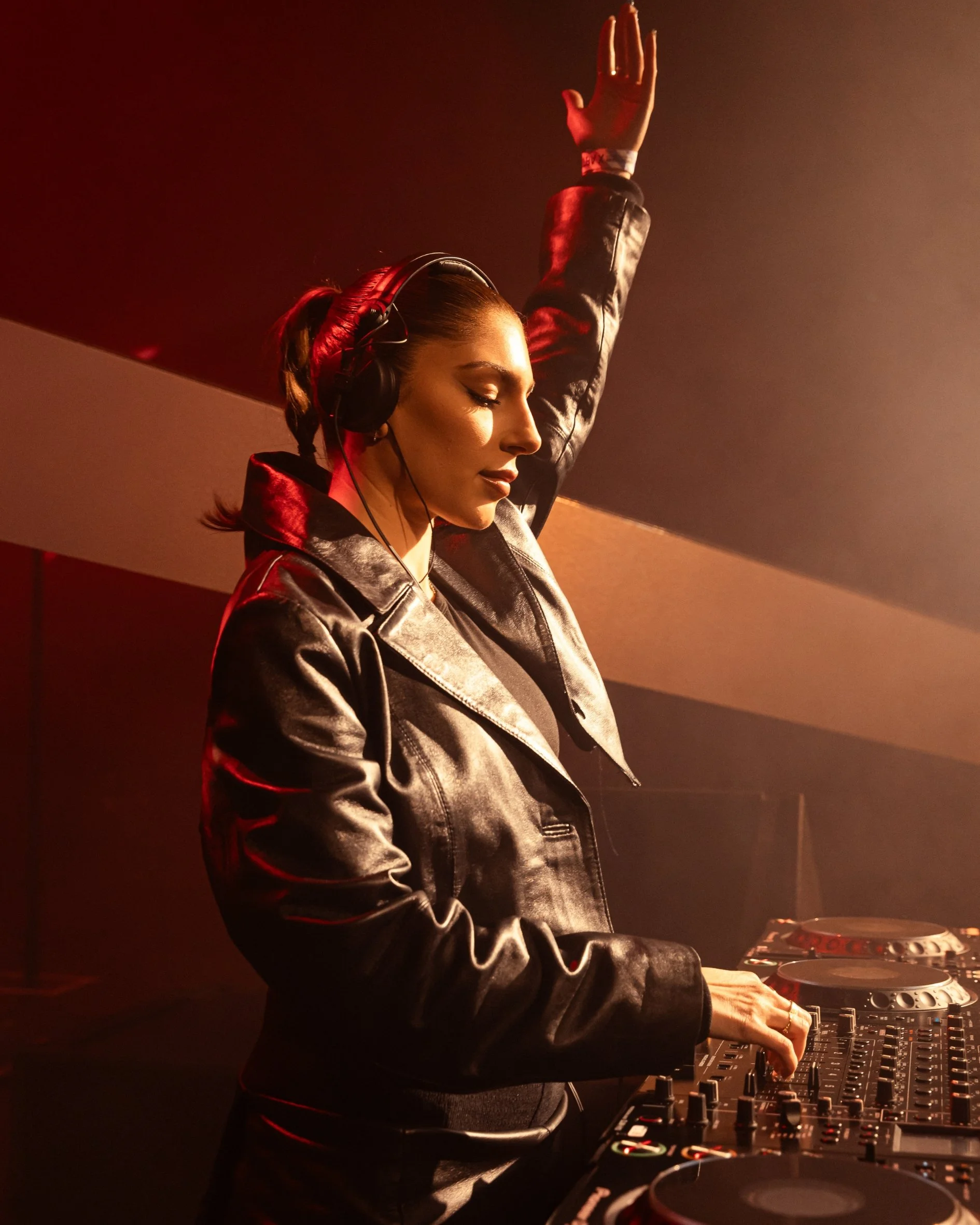 Raphi with a leather jacket and headphones is playing music on DJ equipment in a dimly lit setting with red lighting, raising her right hand with eyes closed.
